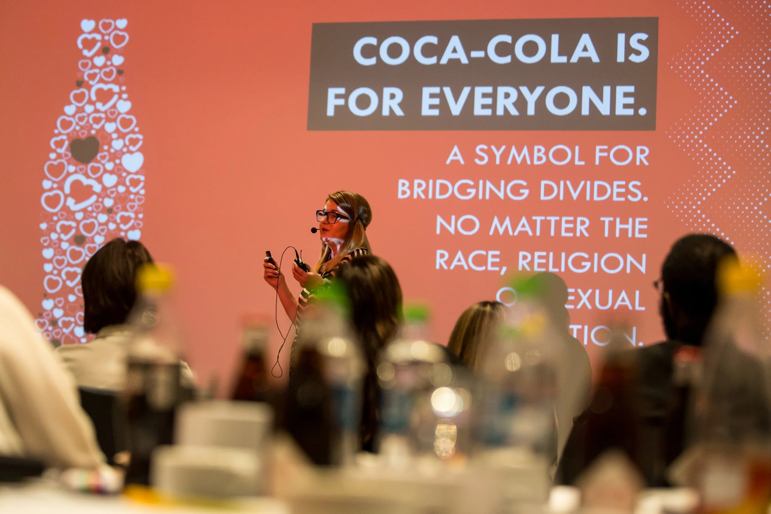 A woman giving a presentation on a stage with a large screen behind her displaying the message 'Coca-Cola is for everyone' and emphasizing the social message that Coca-Cola symbolizes bridging divides regardless of race, religion, sexual orientation.