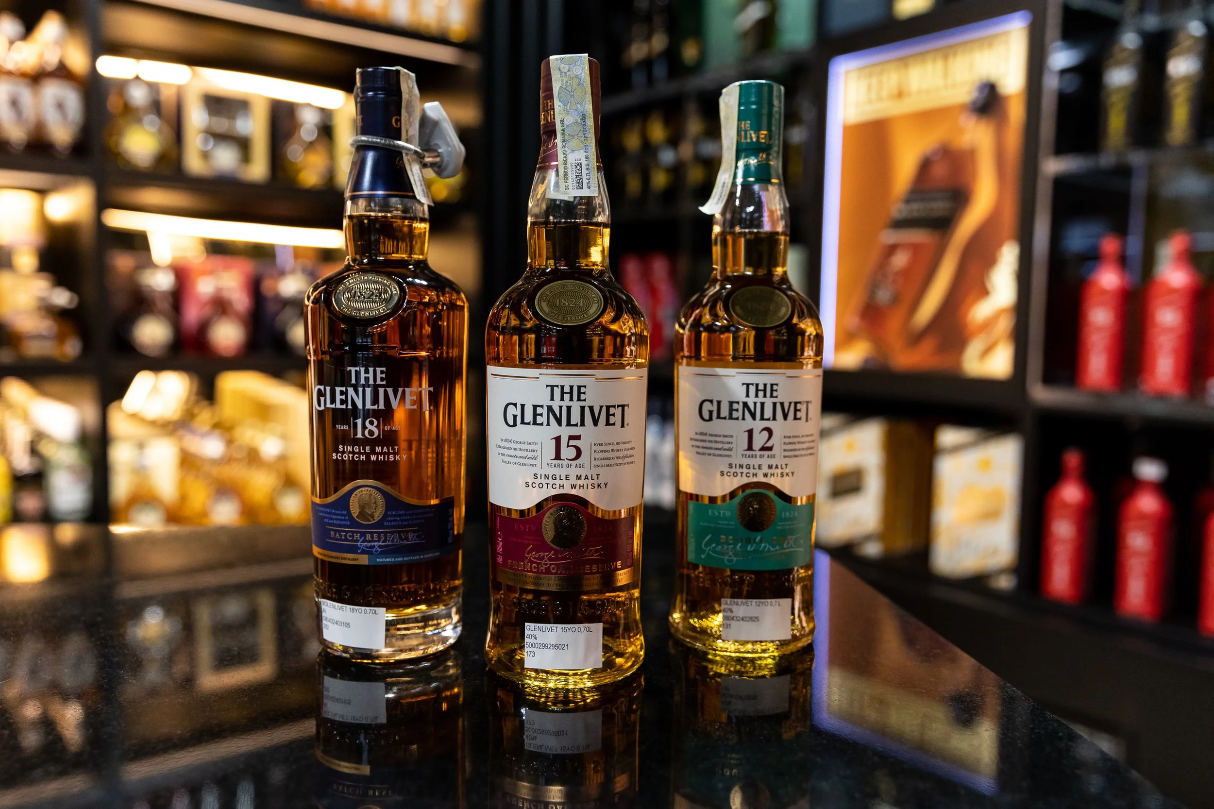 Three bottles of The Glenlivet single malt Scotch whisky on a shelf in a store. The bottles are labeled 18, 15, and 12 years of age. The background is blurred with other bottles and products on shelves.