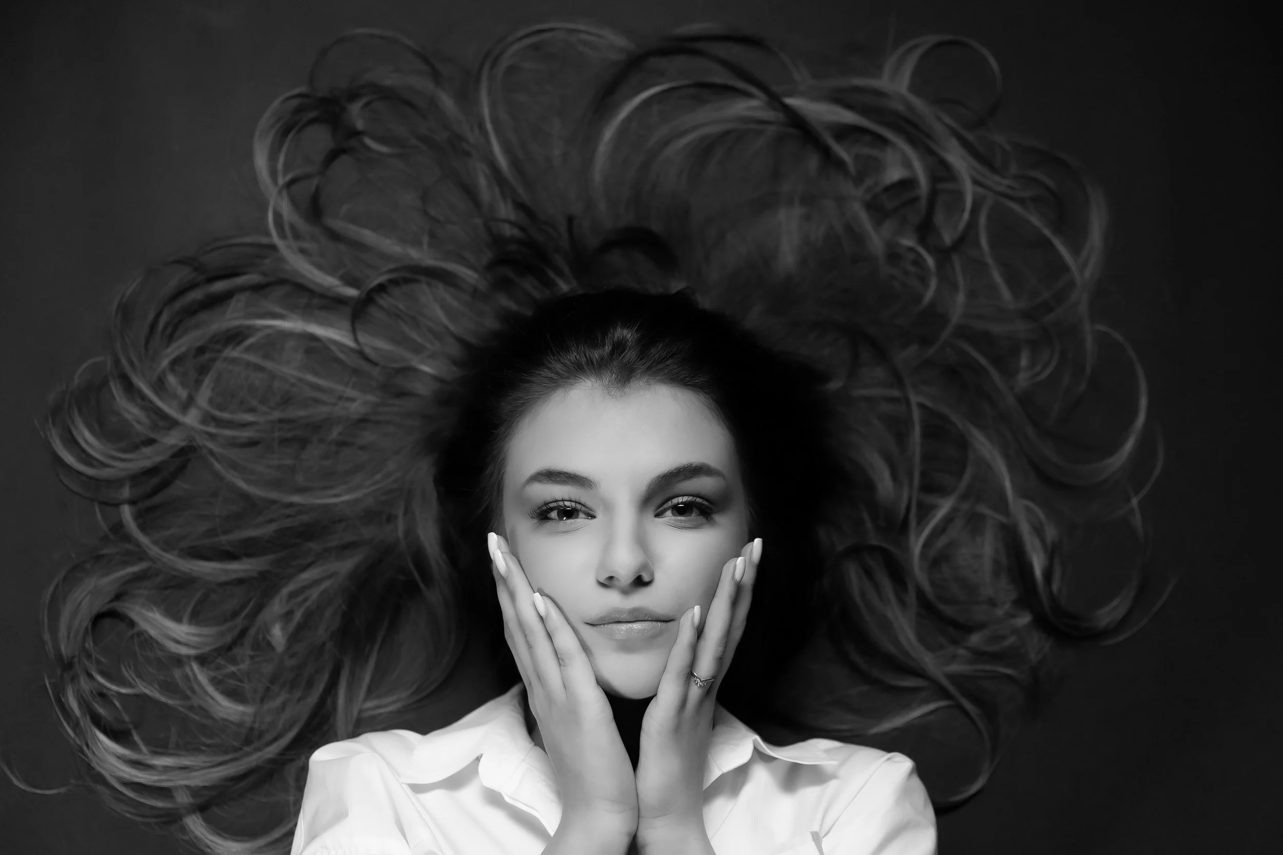 Black and white photo of a woman lying on her back with her long, wavy hair spread around her. She is touching her face with both hands and looking directly at the camera.