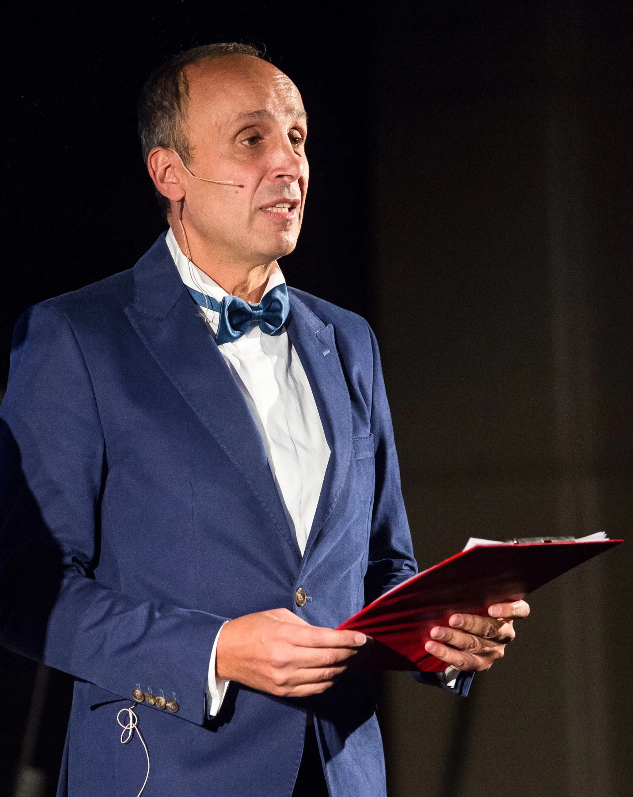 A man wearing a blue tuxedo with a bow tie, holding a red folder, speaking into a headset microphone, on a dark stage.