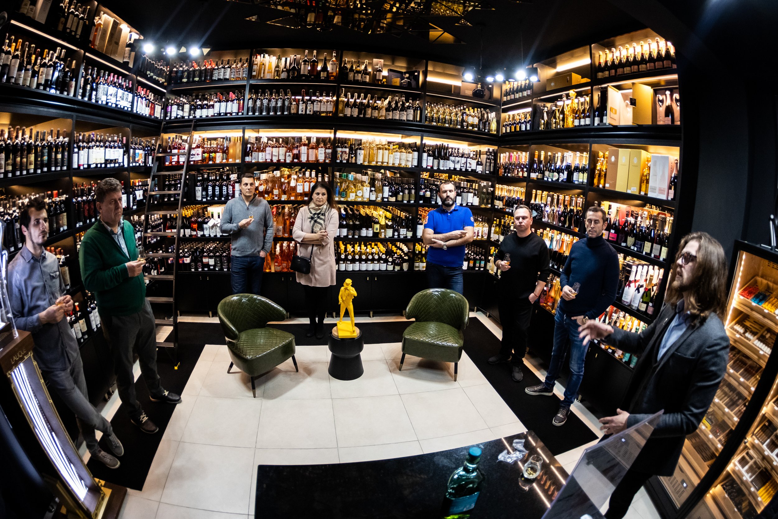 People gathered inside a wine shop, standing around a large wine display with racks full of various bottles. Some are holding wine glasses, and there is a small table with chairs in the center. The shop has dark walls with warm lighting highlighting 