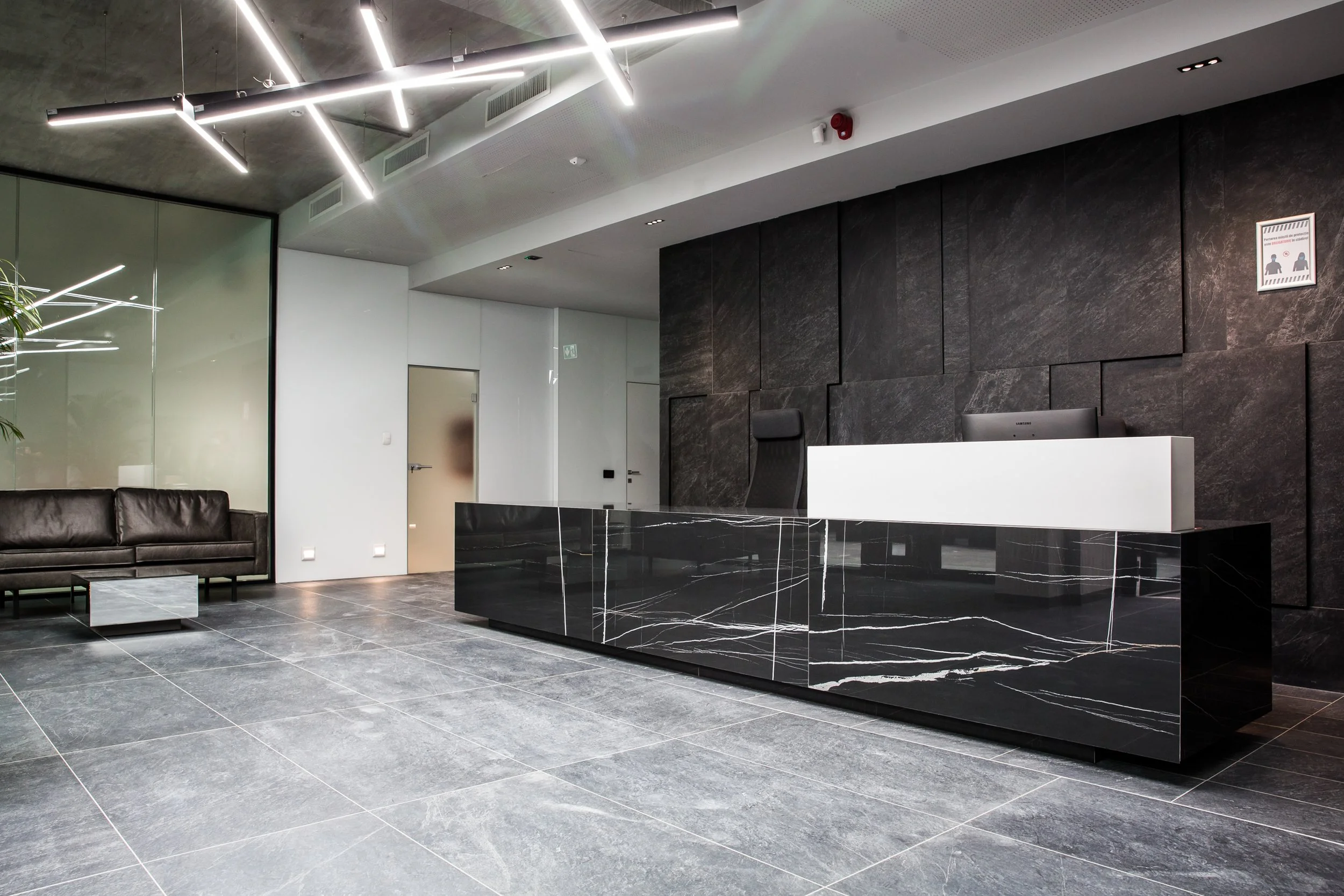 Modern hotel lobby with black marble reception desk, dark gray tiled floor, black textured wall, white and glass accents, leather seating, contemporary lighting, and a sign indicating COVID-19 safety measures.