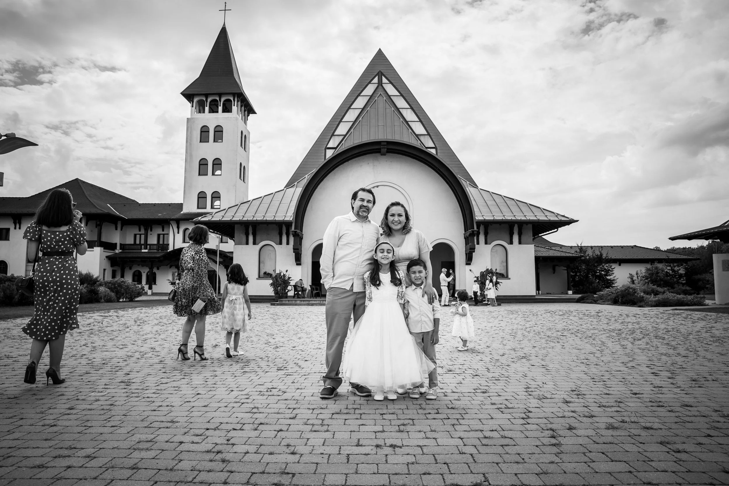 A group of people gathered in front of a church, with a man, woman, and two children smiling in the center. Other small groups of people are visible on either side, with some children, and a woman wearing a polka dot dress taking a photo. The church 