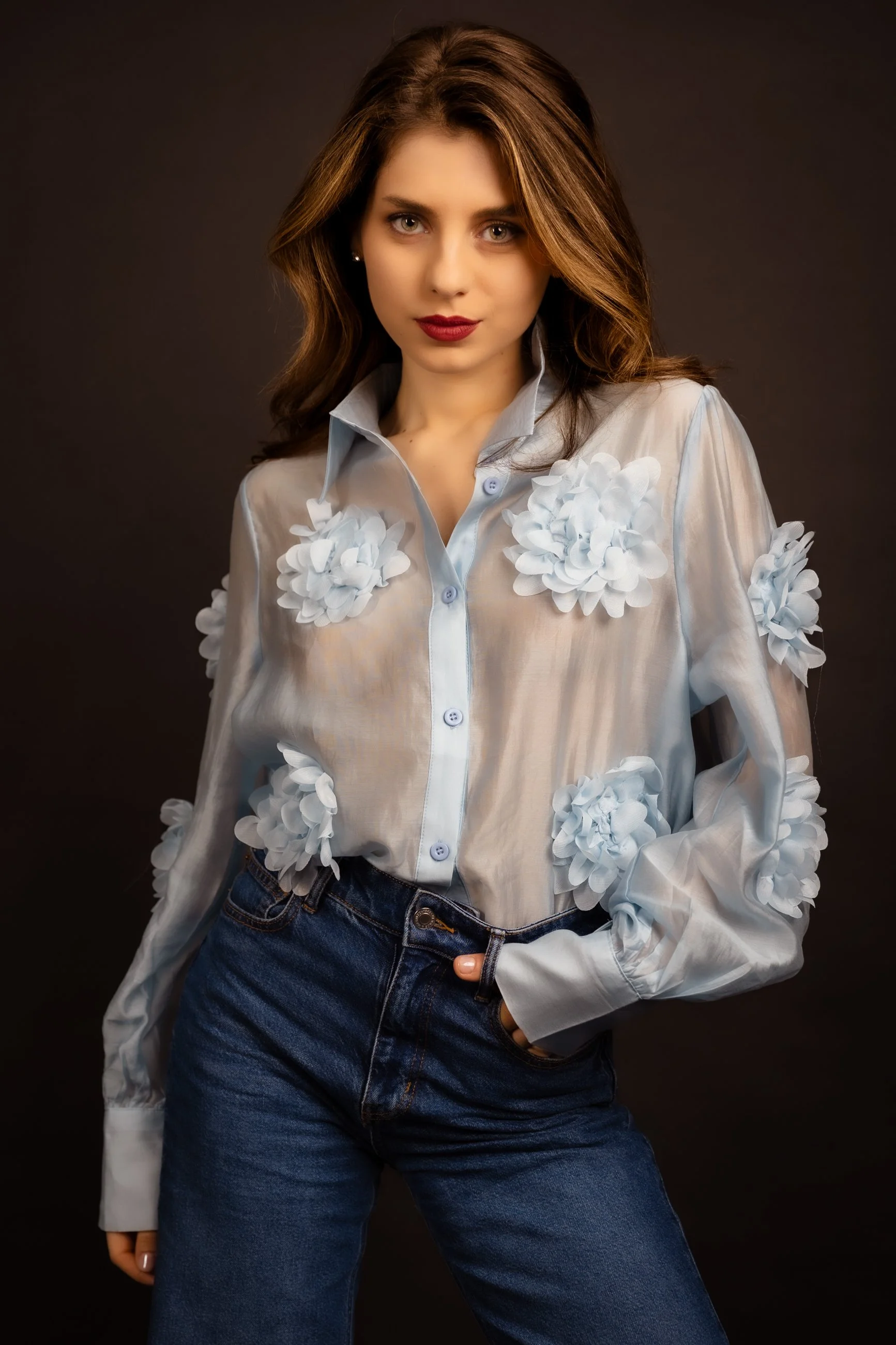 A young woman with brown hair styled in loose waves, wearing a sheer white button-up shirt decorated with white 3D floral appliqués on the shoulders and chest, paired with high-waisted blue jeans, standing against a dark background.