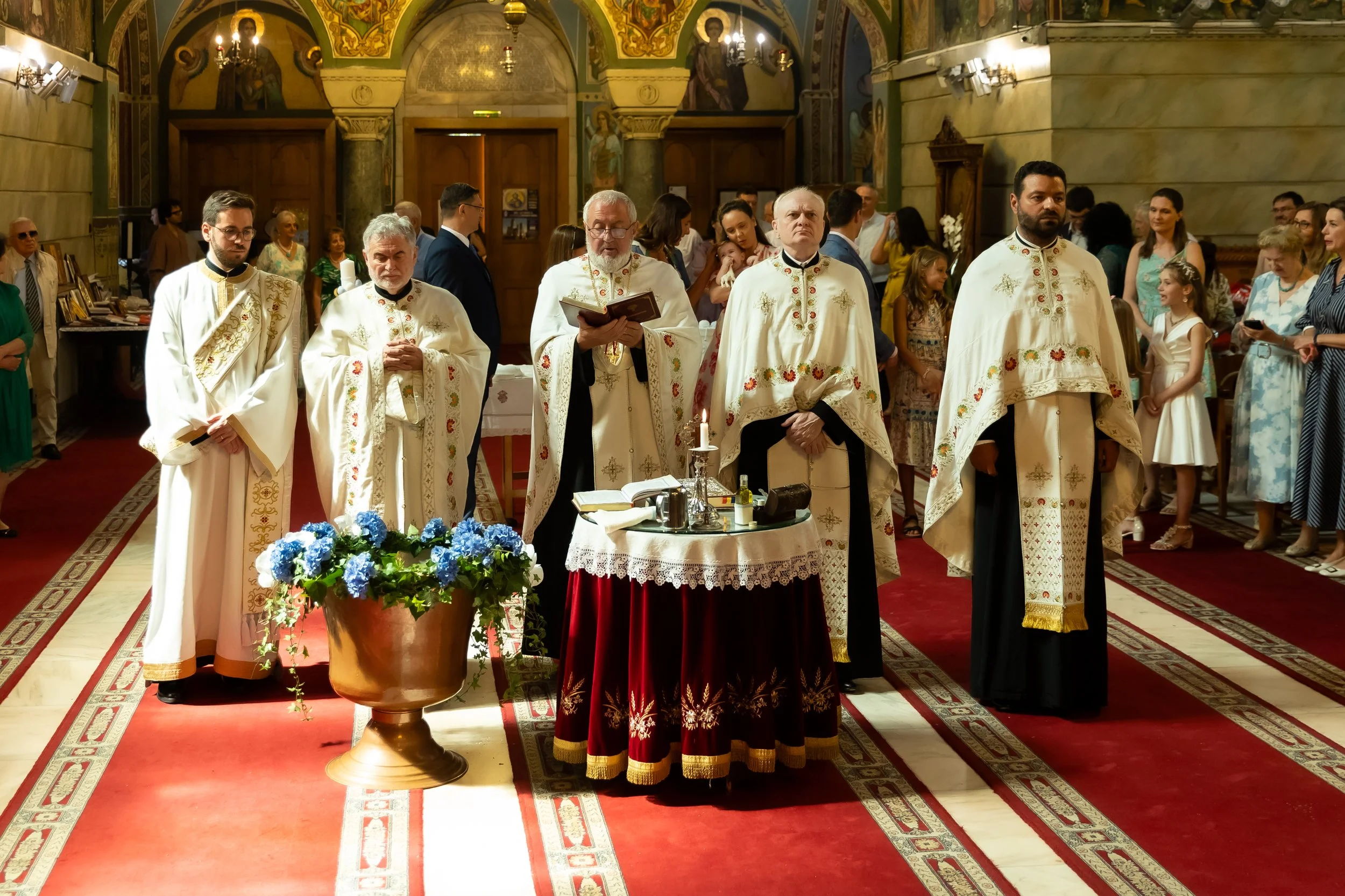 Clergy performing a religious ceremony in a church with ornate iconography and hearlds, various laypeople in attendance.
