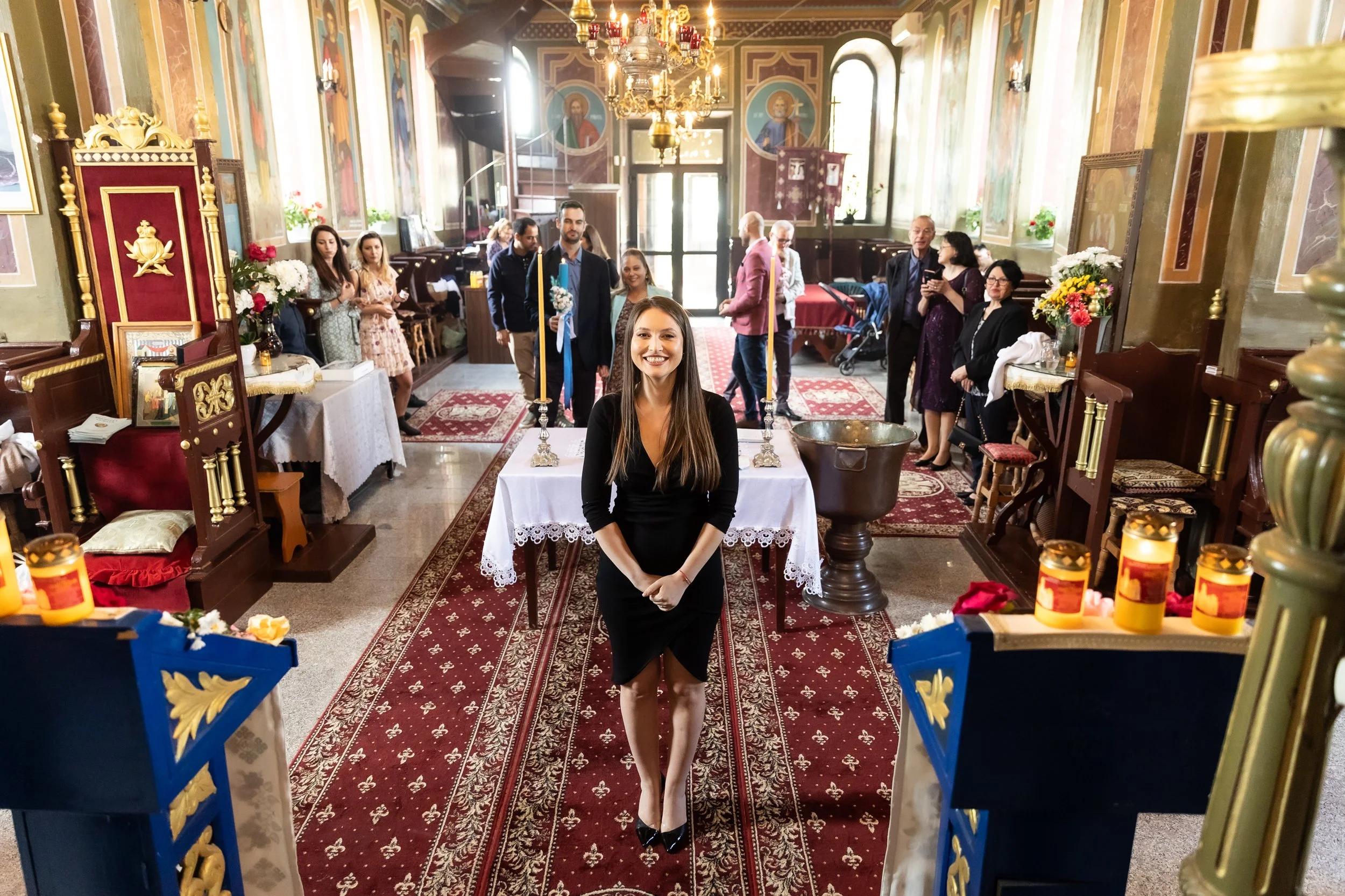 A woman with long brown hair in a black dress is smiling and sitting at a table inside a church, with people gathered in the background.