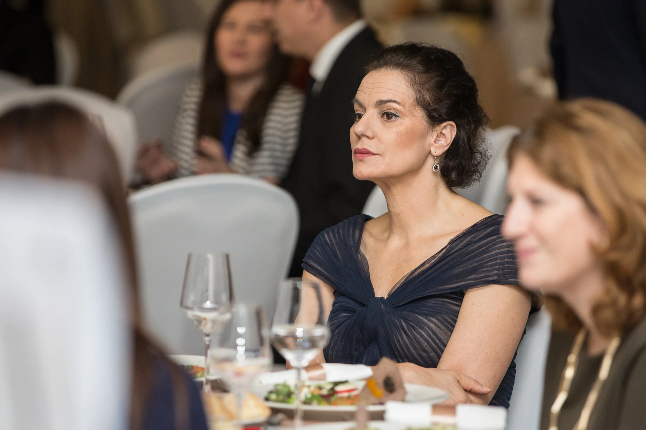 A woman with dark hair and blue earrings sitting at a formal event with glasses of water and plates of food on the table.