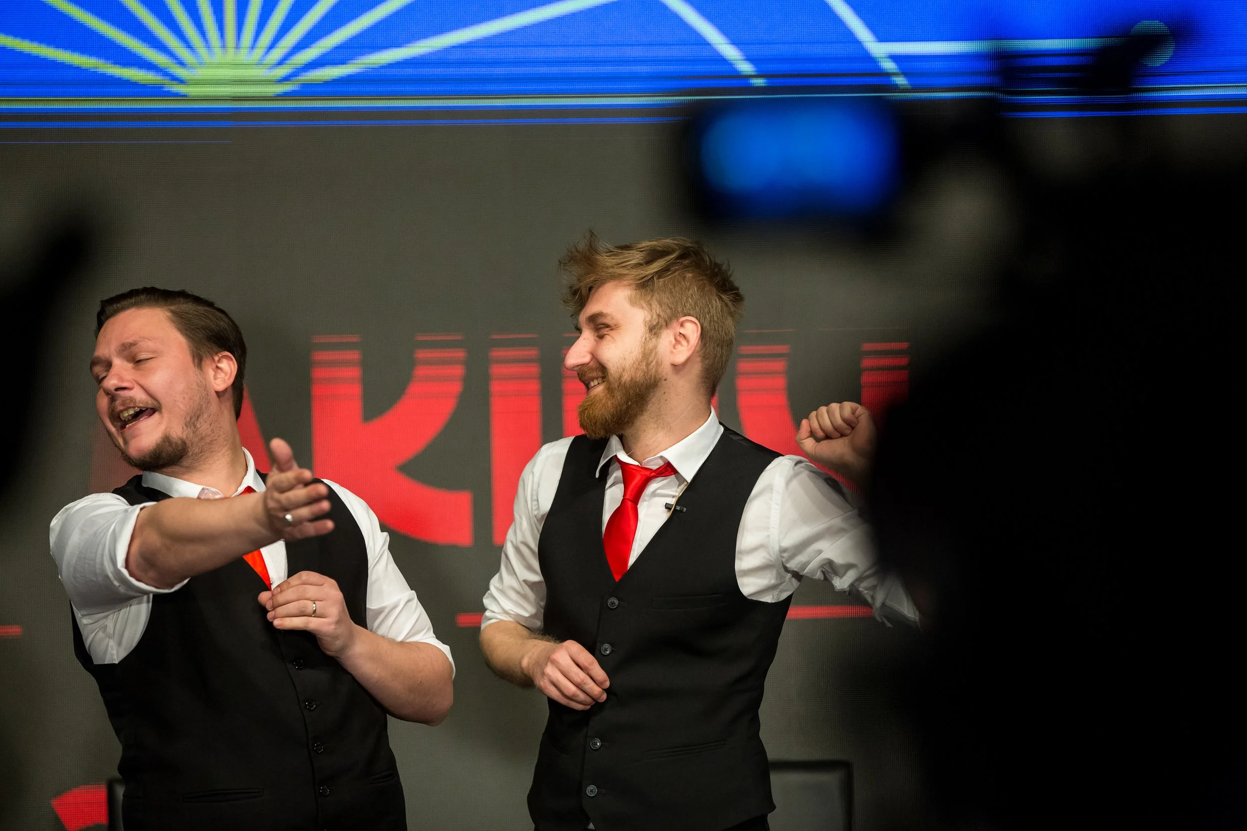 Two men in formal attire celebrating on stage, smiling and making gestures of excitement with a digital screen with red and blue graphics in the background.