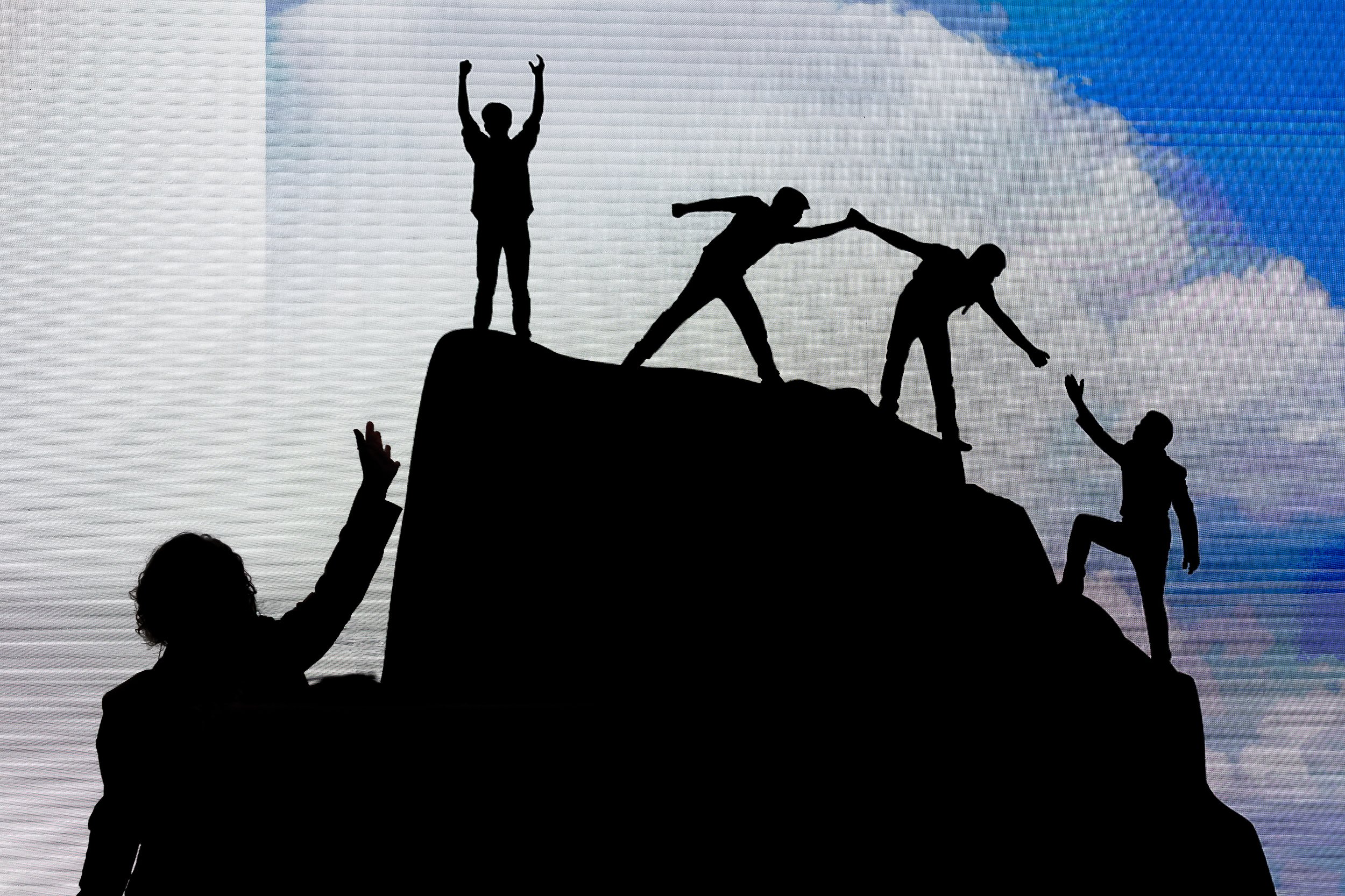 Silhouettes of five people on a mountain peak, with three reaching out for help, one lifting a hand, and one standing with raised arms, against a background of cloudy blue sky.