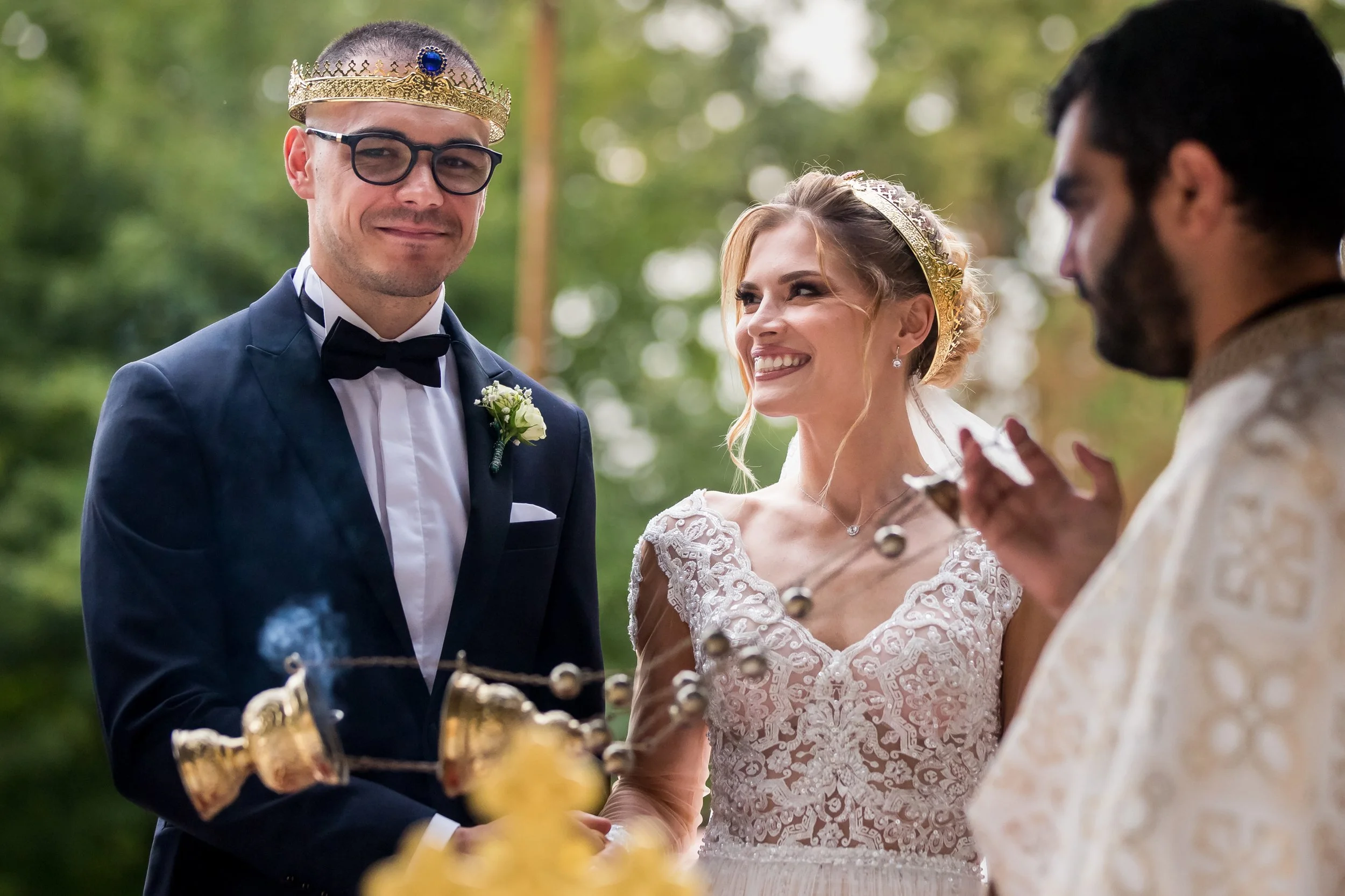 A bride and groom wearing crowns during an outdoor wedding ceremony, holding hands and smiling, with officiant in the background.