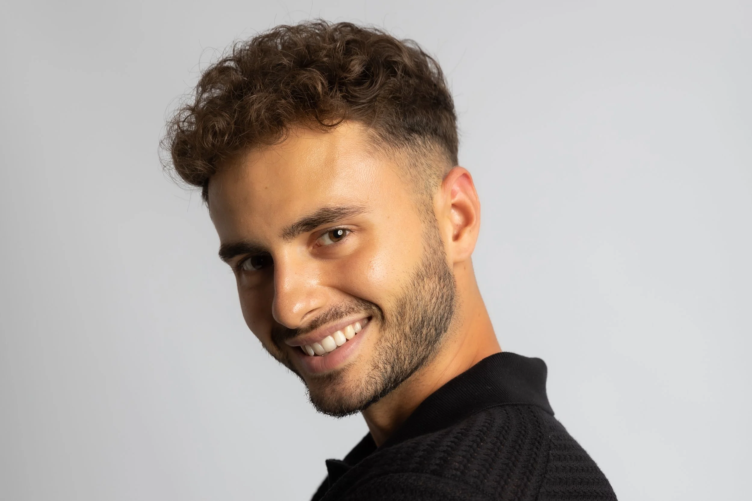 Close-up of a smiling young man with curly brown hair and a beard, wearing a black sweater, against a light gray background.