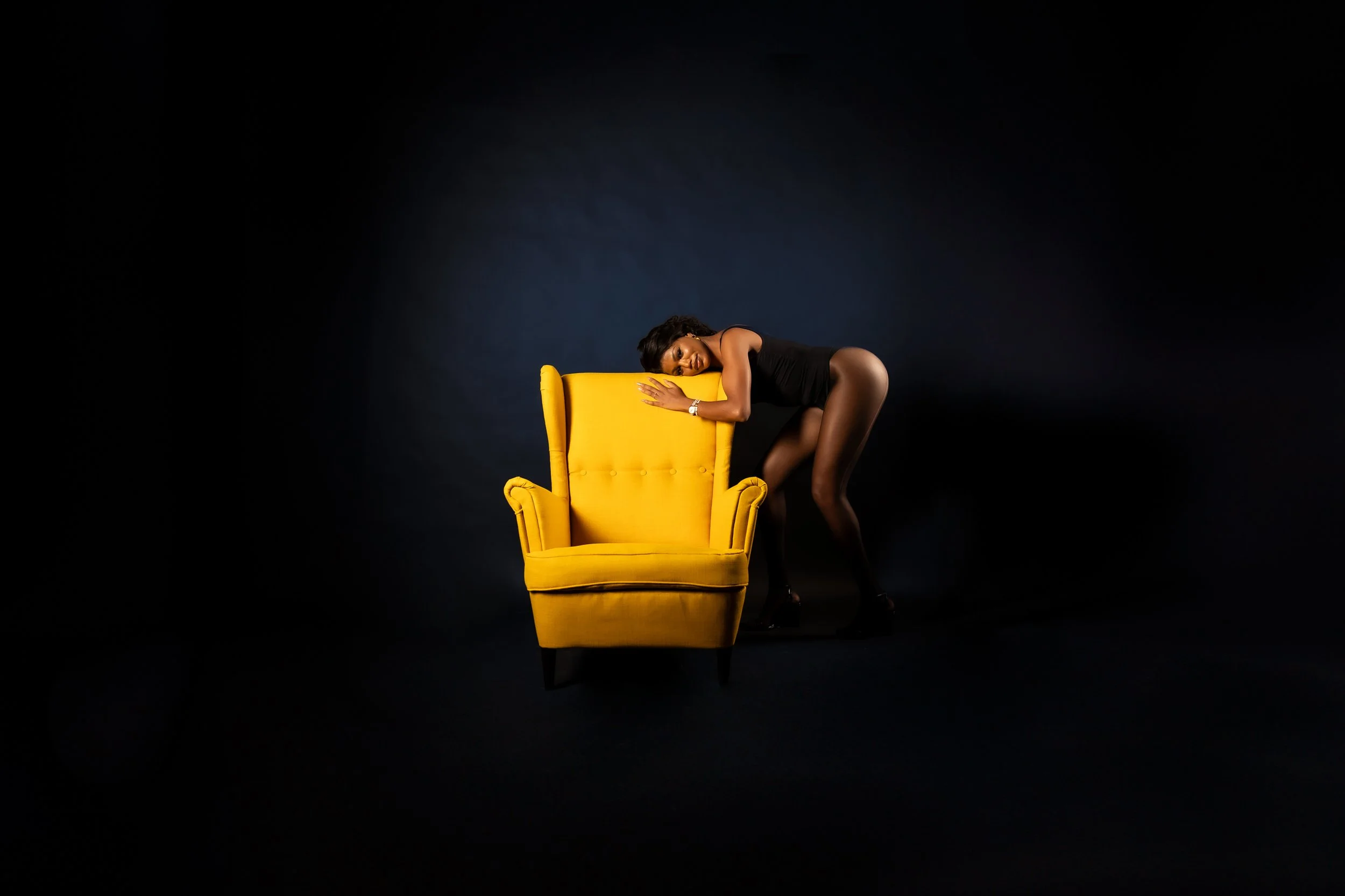 A woman with dark hair in black clothing and high heels is leaning over a bright yellow armchair on a black background.