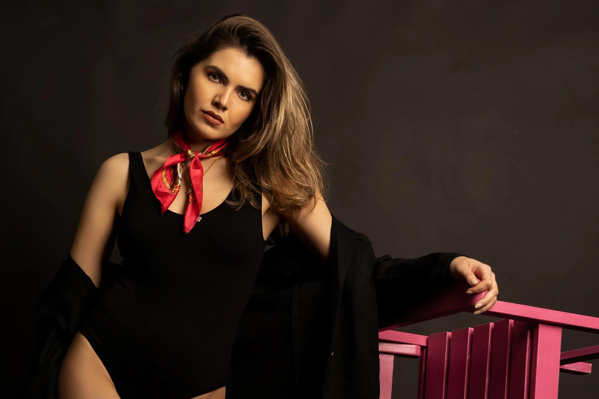 A young woman with long brown hair, wearing a black sleeveless top and a red silk scarf around her neck, poses against a dark background, leaning on a pink wooden railing.