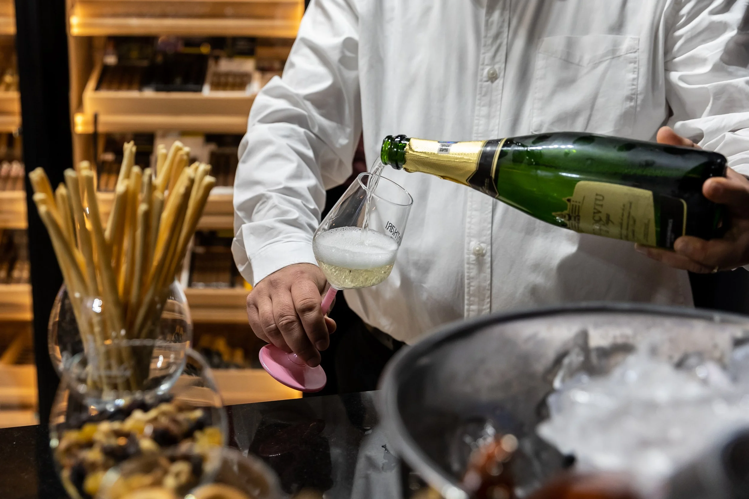 Person in a white shirt pouring champagne into a glass in a cozy, wooden-shelved setting.