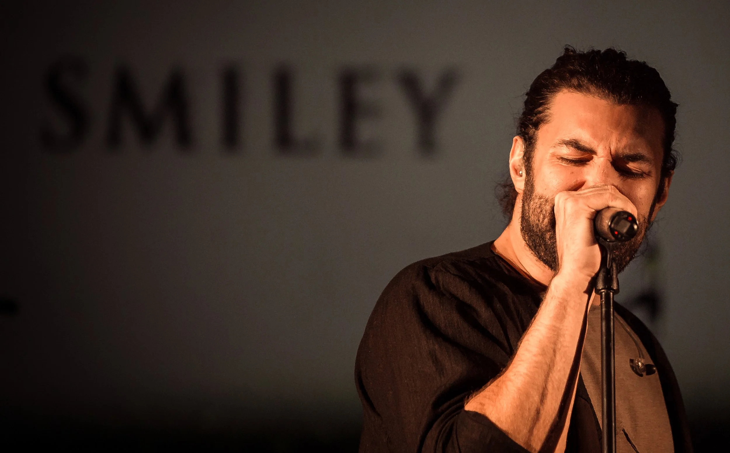 A man with curly hair and a beard sings into a microphone with his eyes closed, wearing a black shirt, in a dimly lit setting.