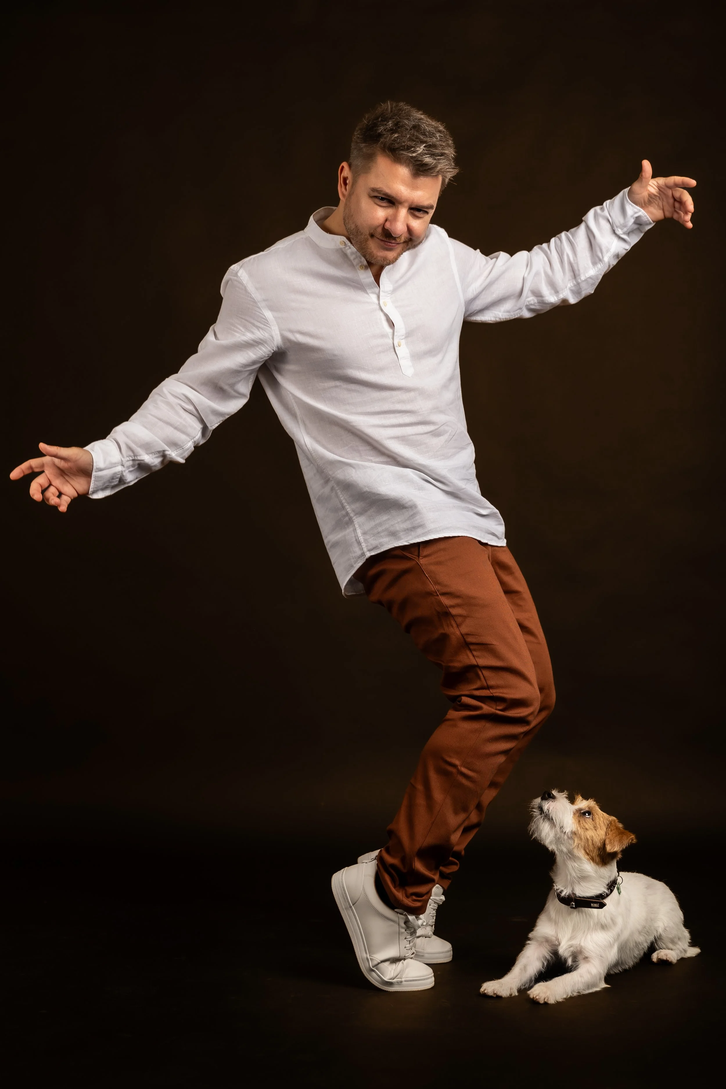 A man in a white shirt and brown pants dancing or posing with a dog that is sitting and looking up, on a dark background.