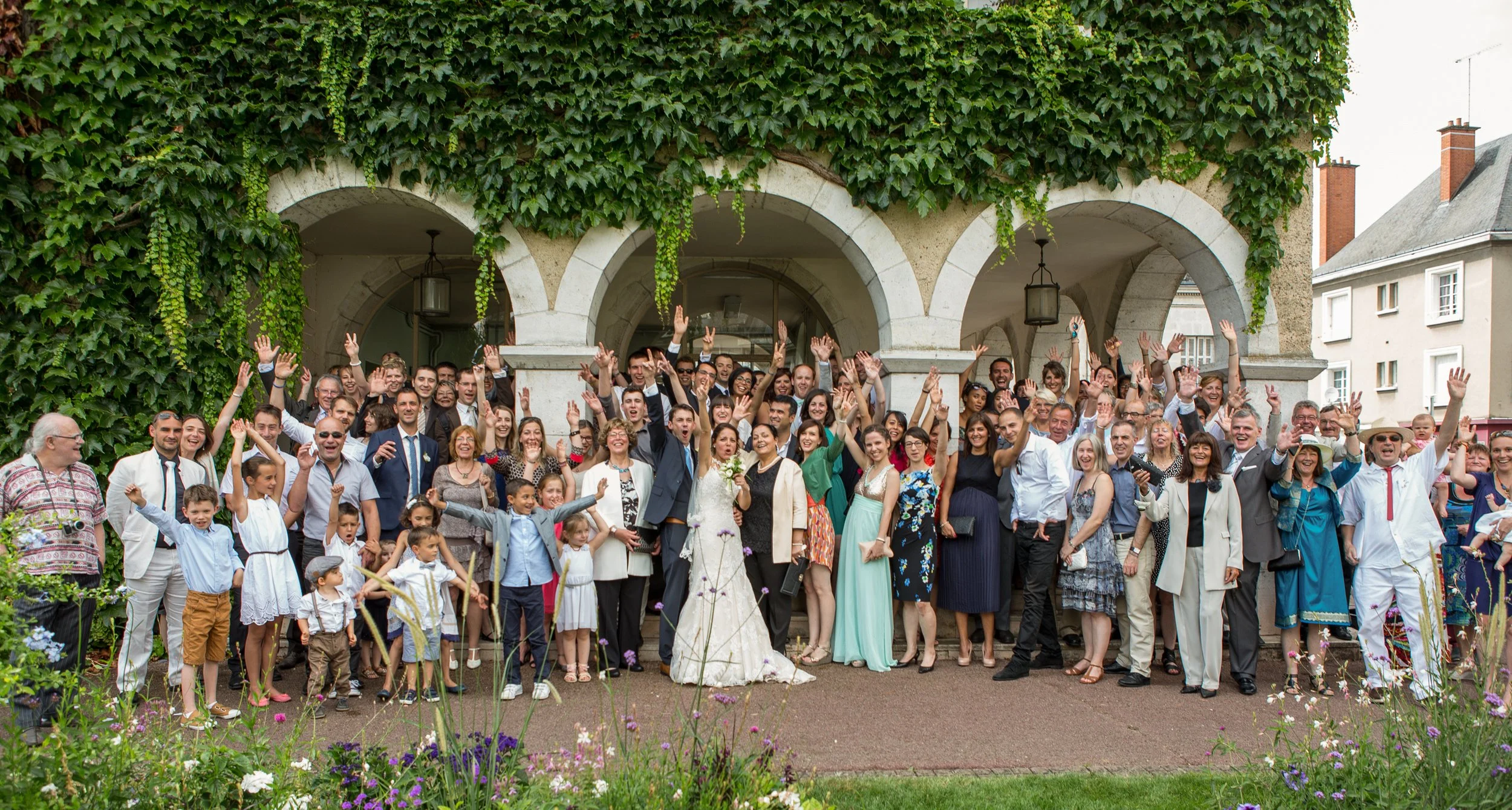 A large group of people celebrating outdoors, many raising their hands, in front of a building covered in green ivy and plants, during daytime.