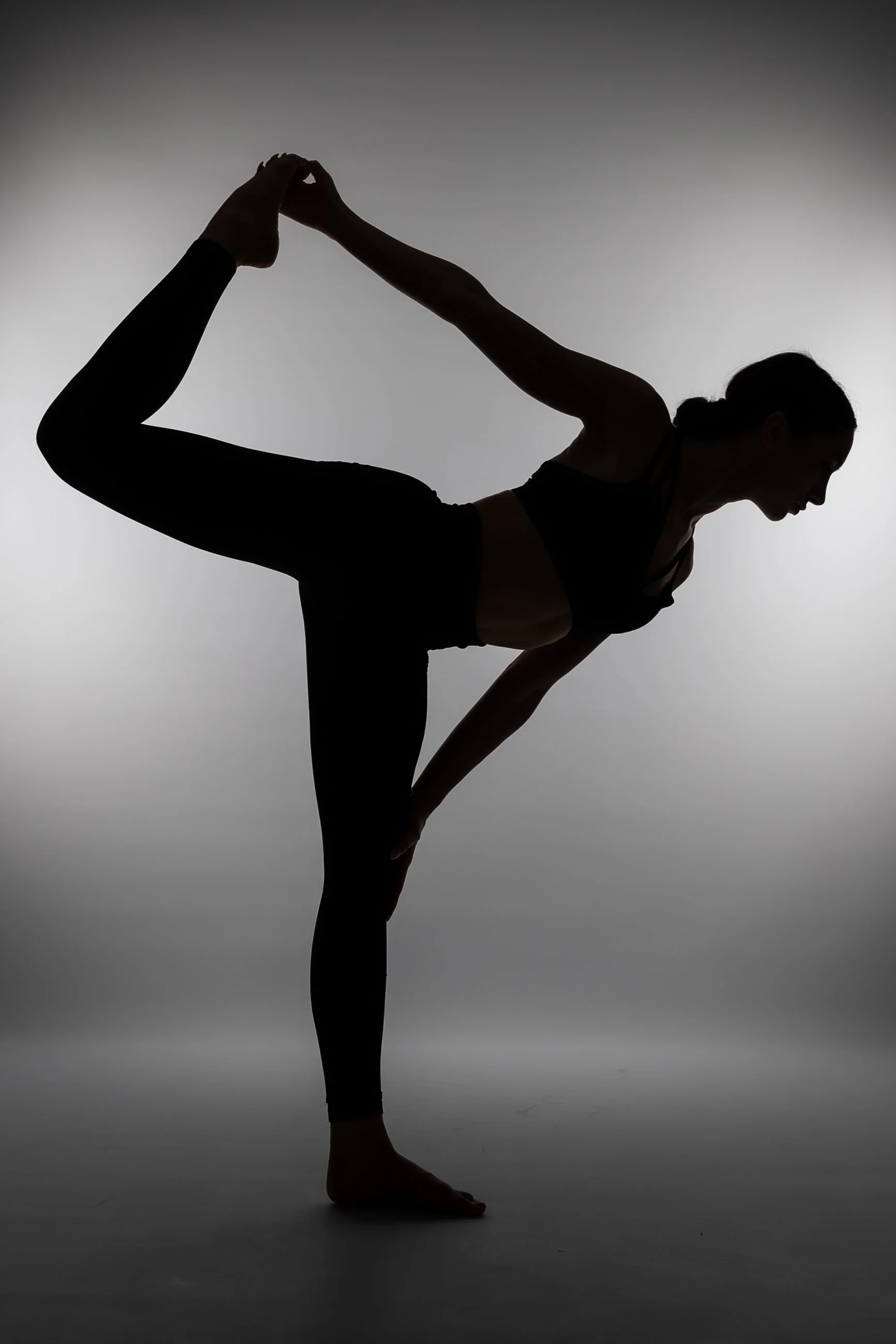 Silhouette of a woman performing a yoga pose with one leg bent and lifted, holding her foot with her hand, against a plain gray background.