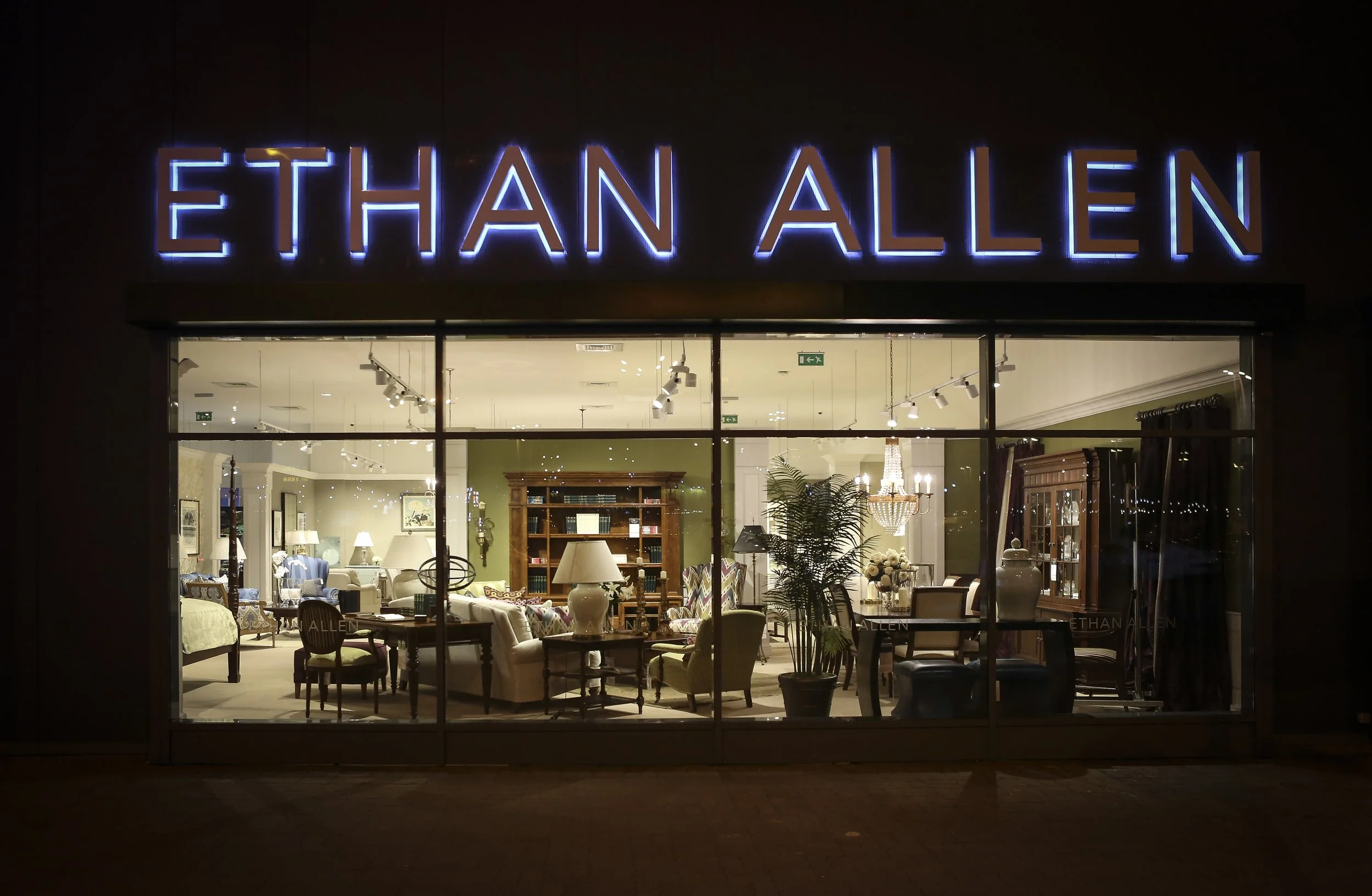 Night view of Ethan Allen furniture store with brightly lit interior showcasing furniture and home decor items.