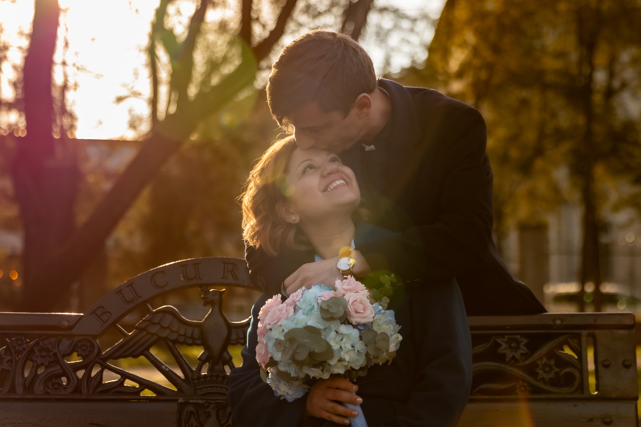 A young woman sitting on a park bench holding a bouquet of pink and white flowers while a young man leans over her, touching her forehead with his nose. The scene is set outdoors during golden hour, with sunlight filtering through the trees in the ba