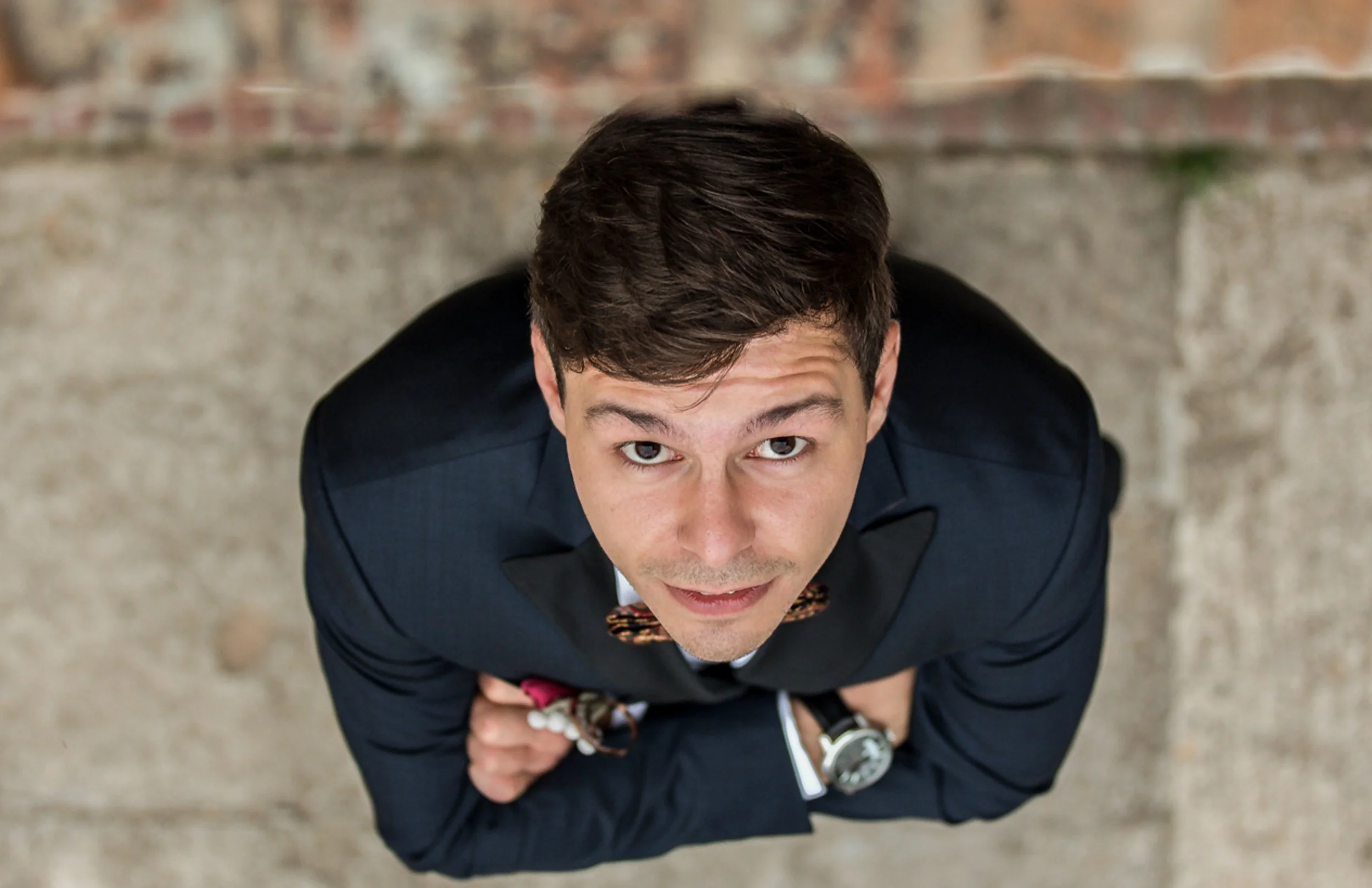 A young man in a suit and bow tie, looking up at the camera, standing outdoors on a concrete surface with a brick wall background.
