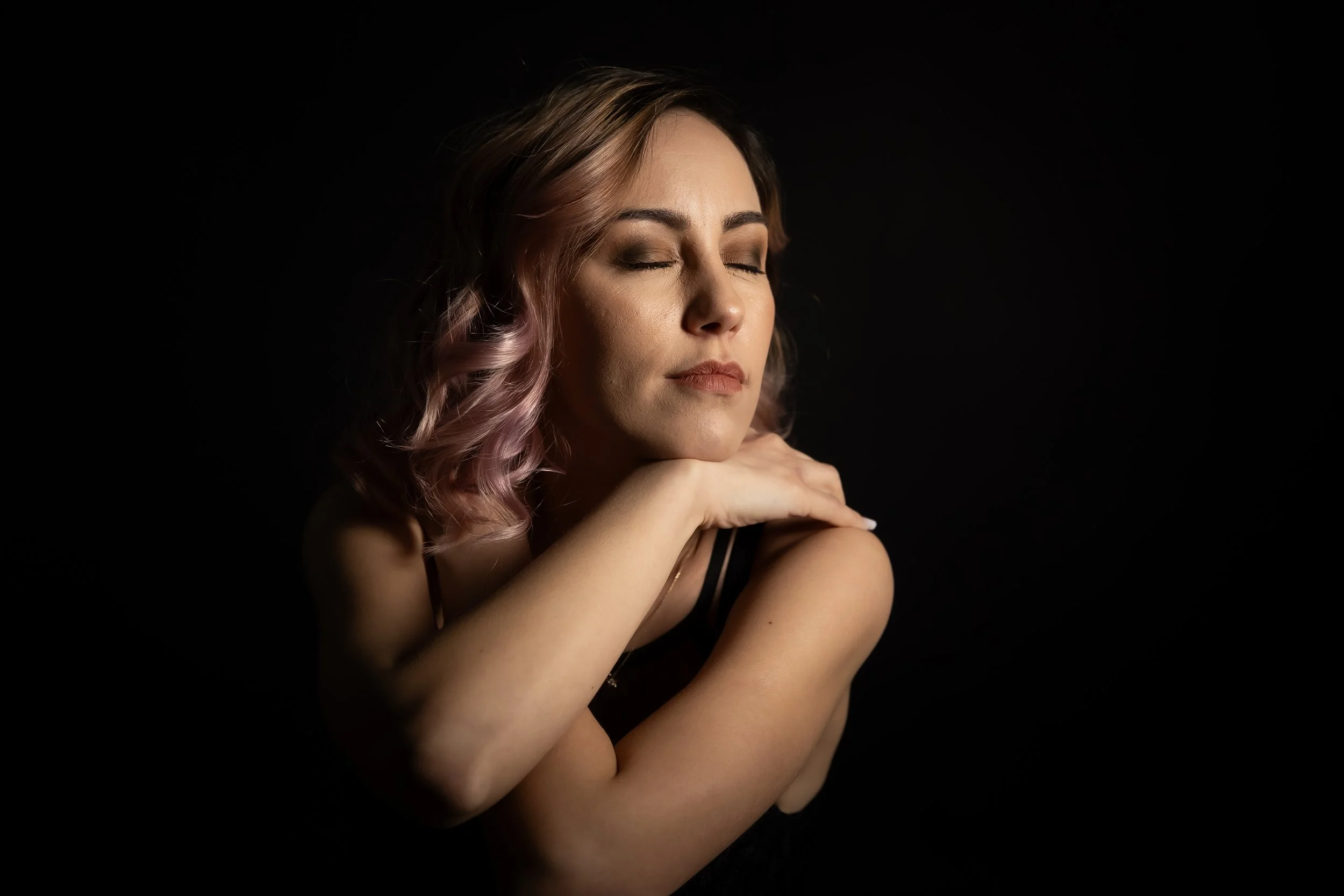 A woman with closed eyes and wavy hair with pink tips, resting her chin on her crossed arms, against a black background.