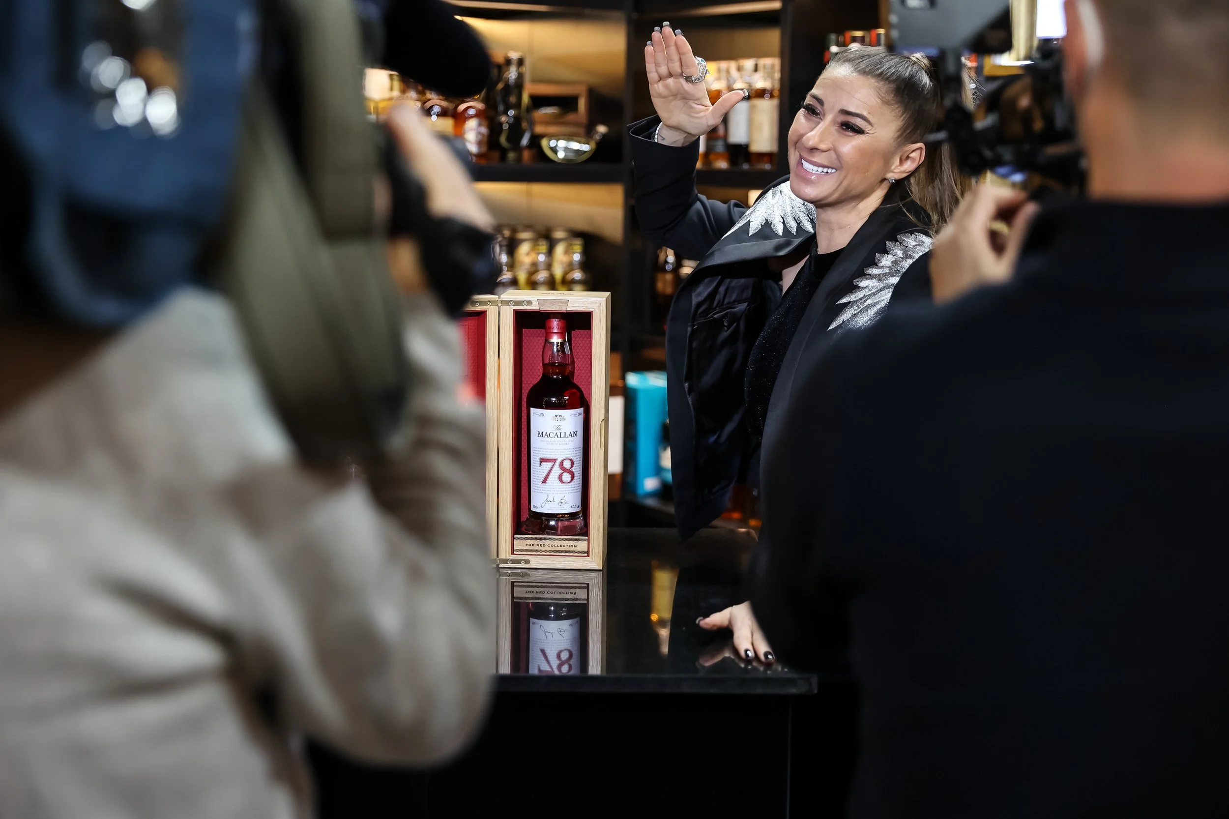 Woman smiling and waving while being recorded by a camera in a liquor store, with a bottle of whiskey displayed on the counter.