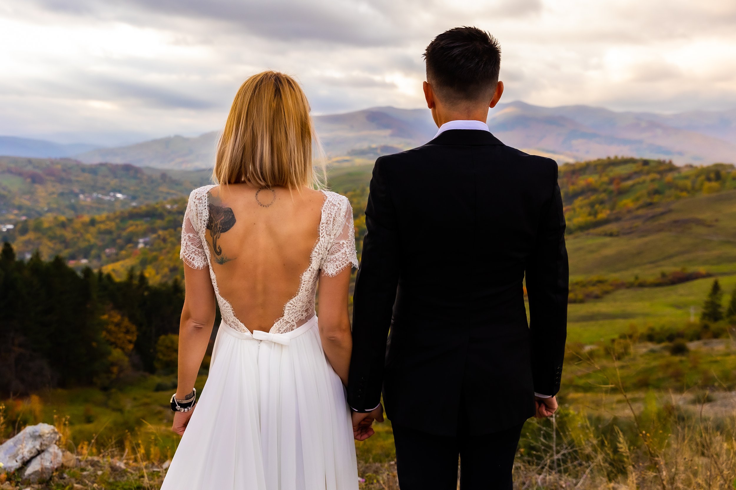 A couple, dressed in wedding attire, holding hands and standing outdoors with a scenic mountainous landscape in the background.