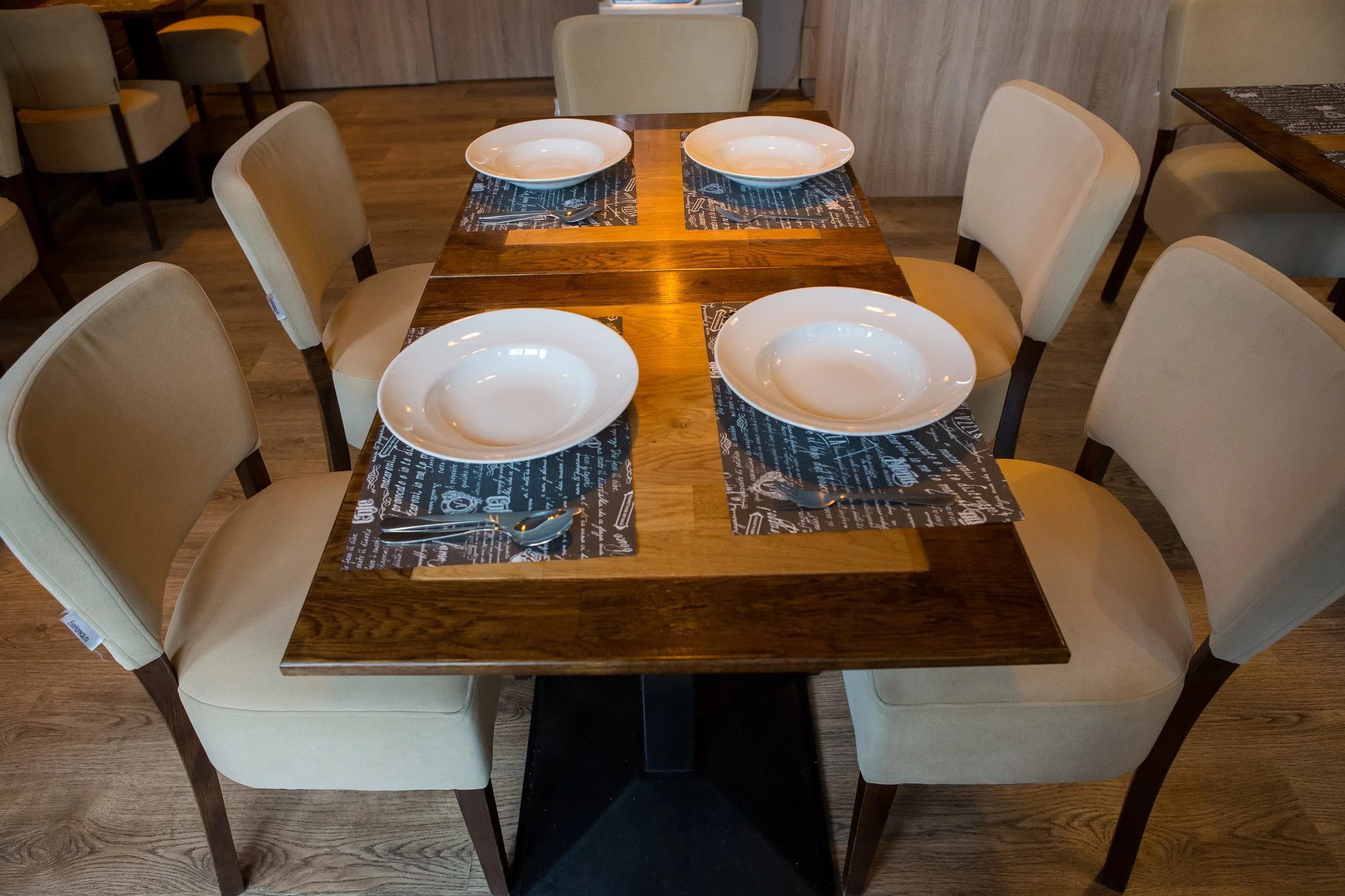 A wooden dining table set for four with white bowls, silverware, and black and white printed placemats in a restaurant or cafe setting.
