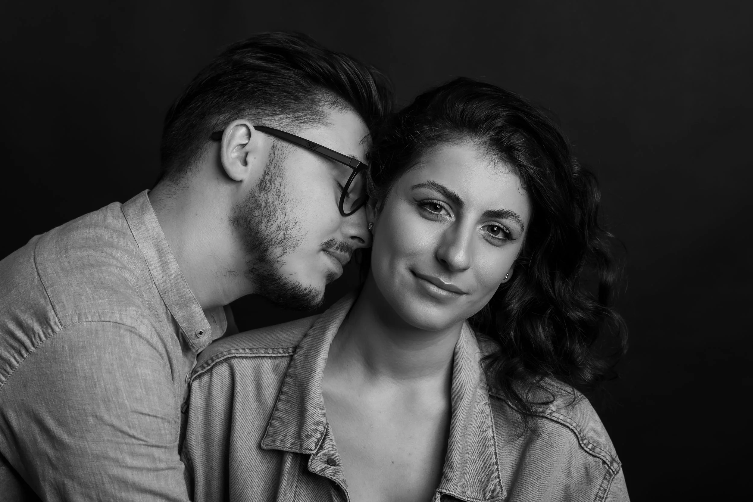 A black and white photo of a man and woman close together, with the man whispering into the woman's ear, both wearing casual clothes.