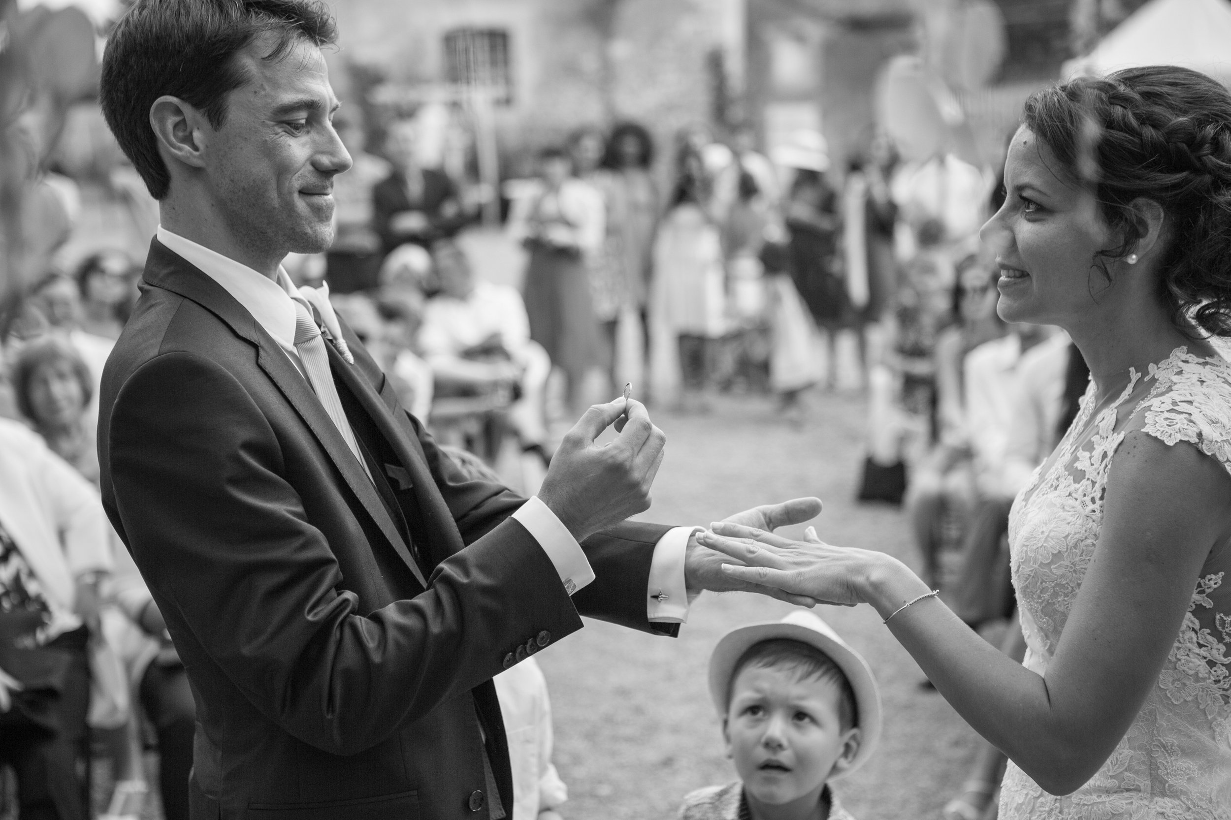 A black and white photo of a wedding ceremony where a man in a suit is placing a ring on a woman's finger. The woman is wearing a lace wedding dress and smiling. There are guests in the background, including a young boy looking up at the couple.