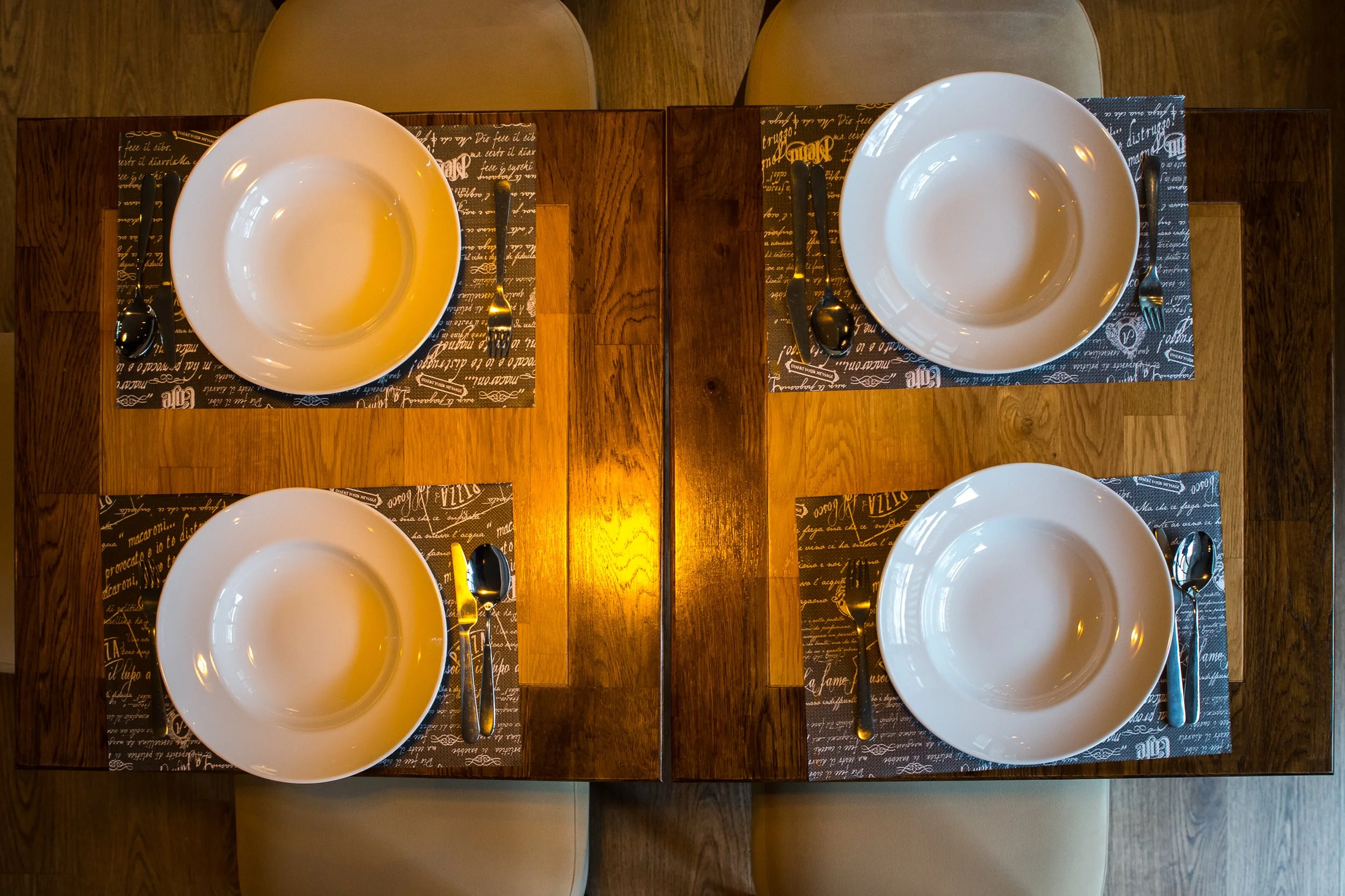 A top-down view of a wooden dining table set for four, with white bowls, black utensils, and black and white patterned placemats.