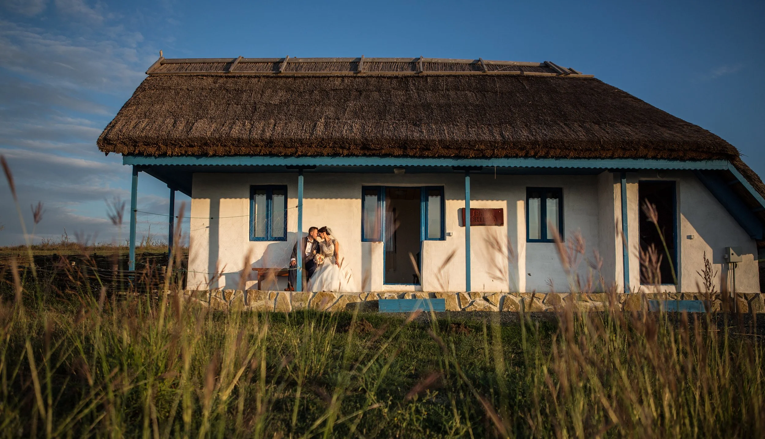 A couple dressed in wedding attire sharing a kiss in front of a small house with a thatched roof during sunset.