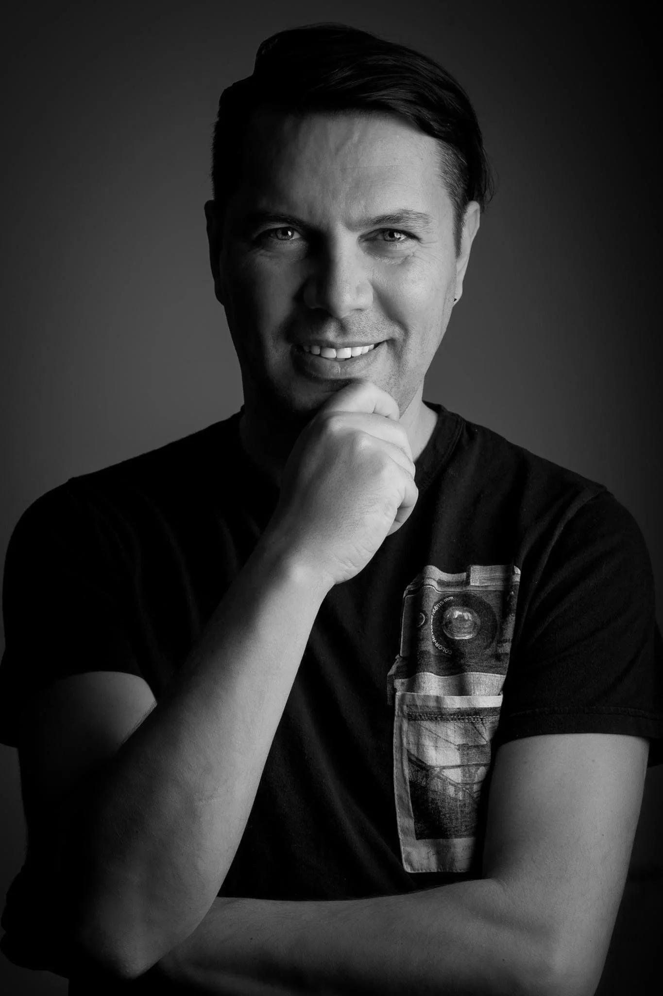 Black and white portrait of a young man with dark hair, smiling, resting his chin on his hand, wearing a T-shirt with a graphic design.
