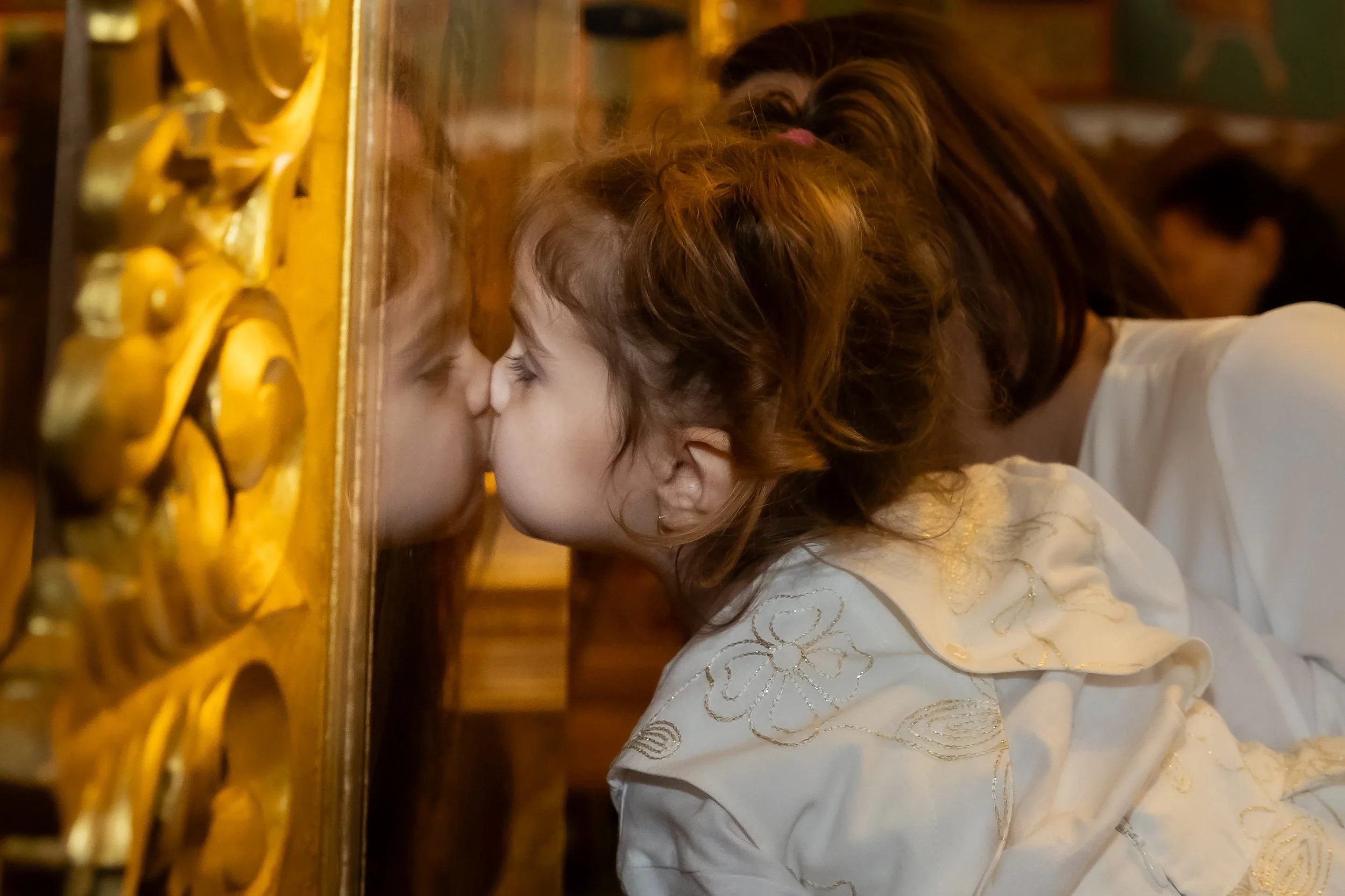 A young girl with curly red hair kissing or resting her face against a wooden surface with ornate gold detailing, possibly a piano or decorative panel, in a warmly lit room.