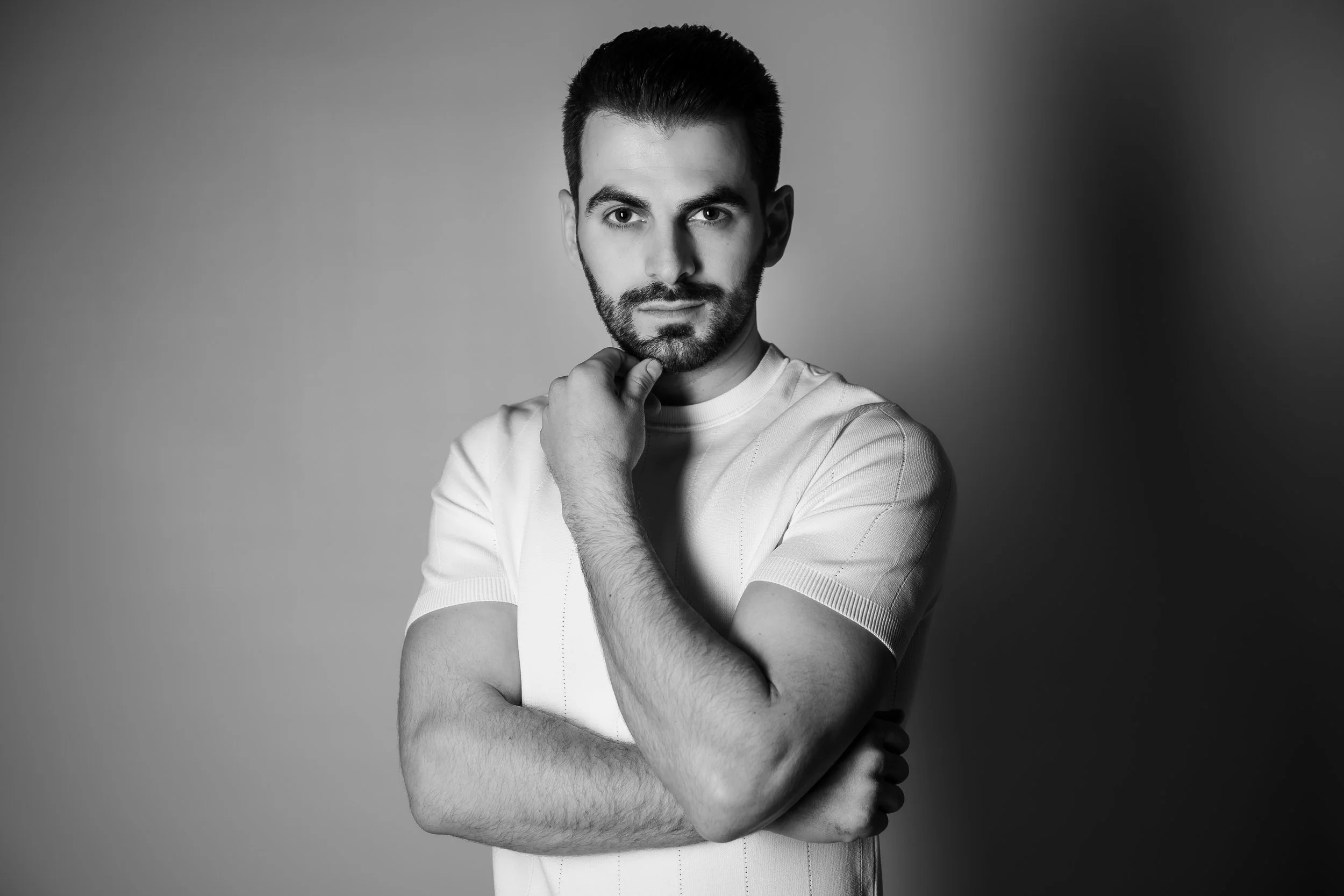 Black and white portrait of a young man with dark hair, beard, and mustache, wearing a light-colored t-shirt, standing against a plain background. He has a serious expression, with one hand touching his chin and the other crossed over his chest.