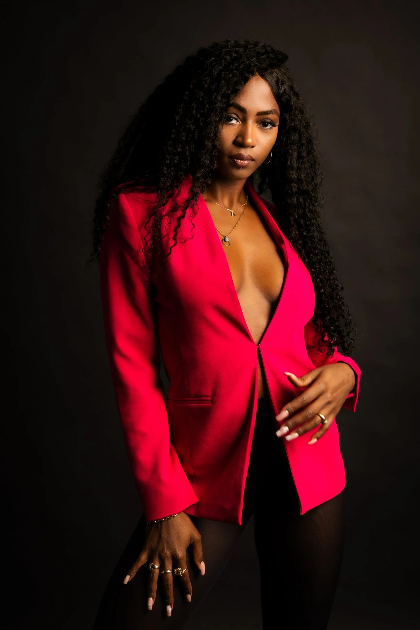 A woman with long curly dark hair wearing a bright pink blazer, black top, and jewelry posing against a dark background.