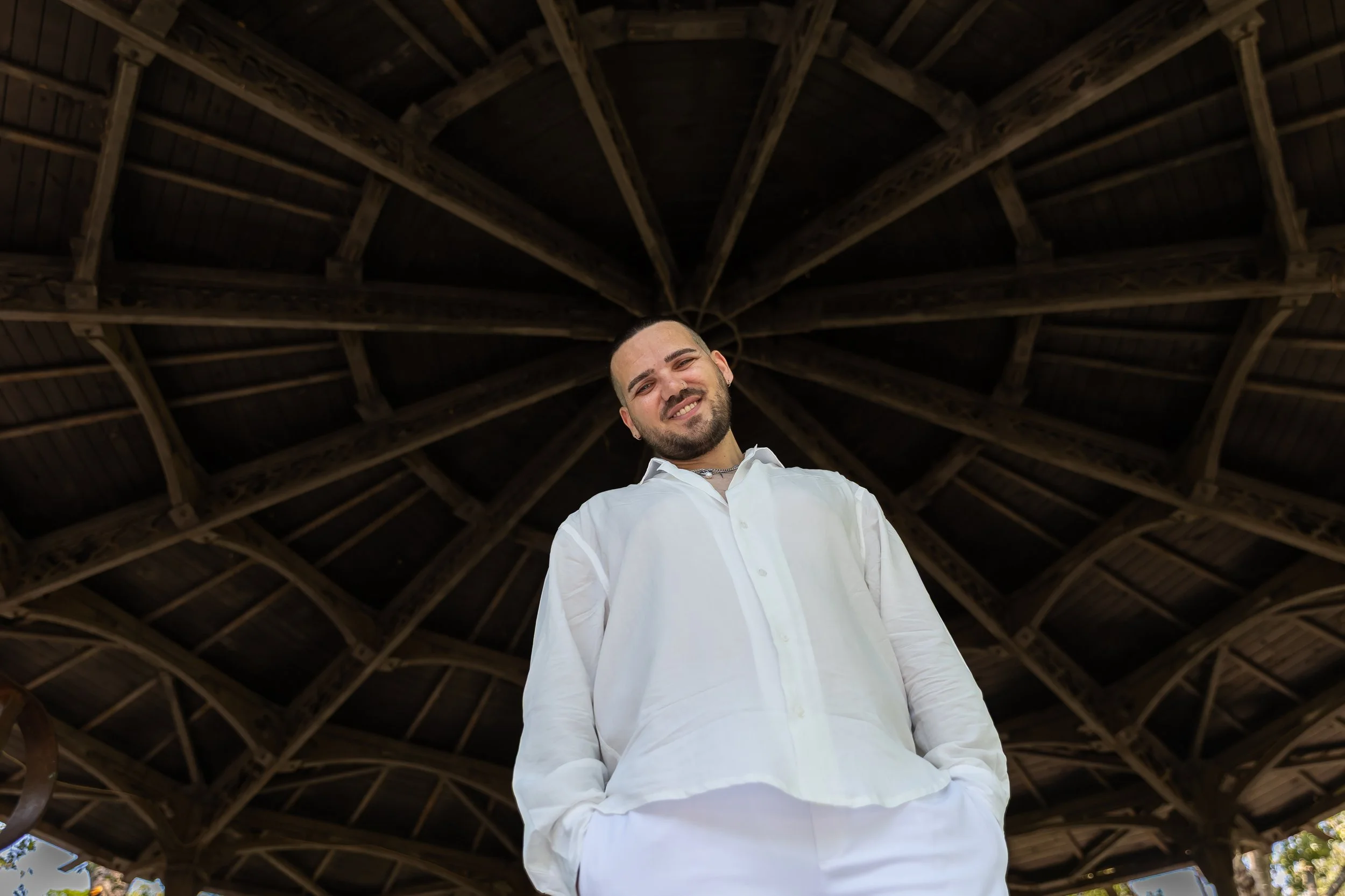A smiling man wearing a white shirt, standing underneath a wooden pavilion with a high, vaulted ceiling.