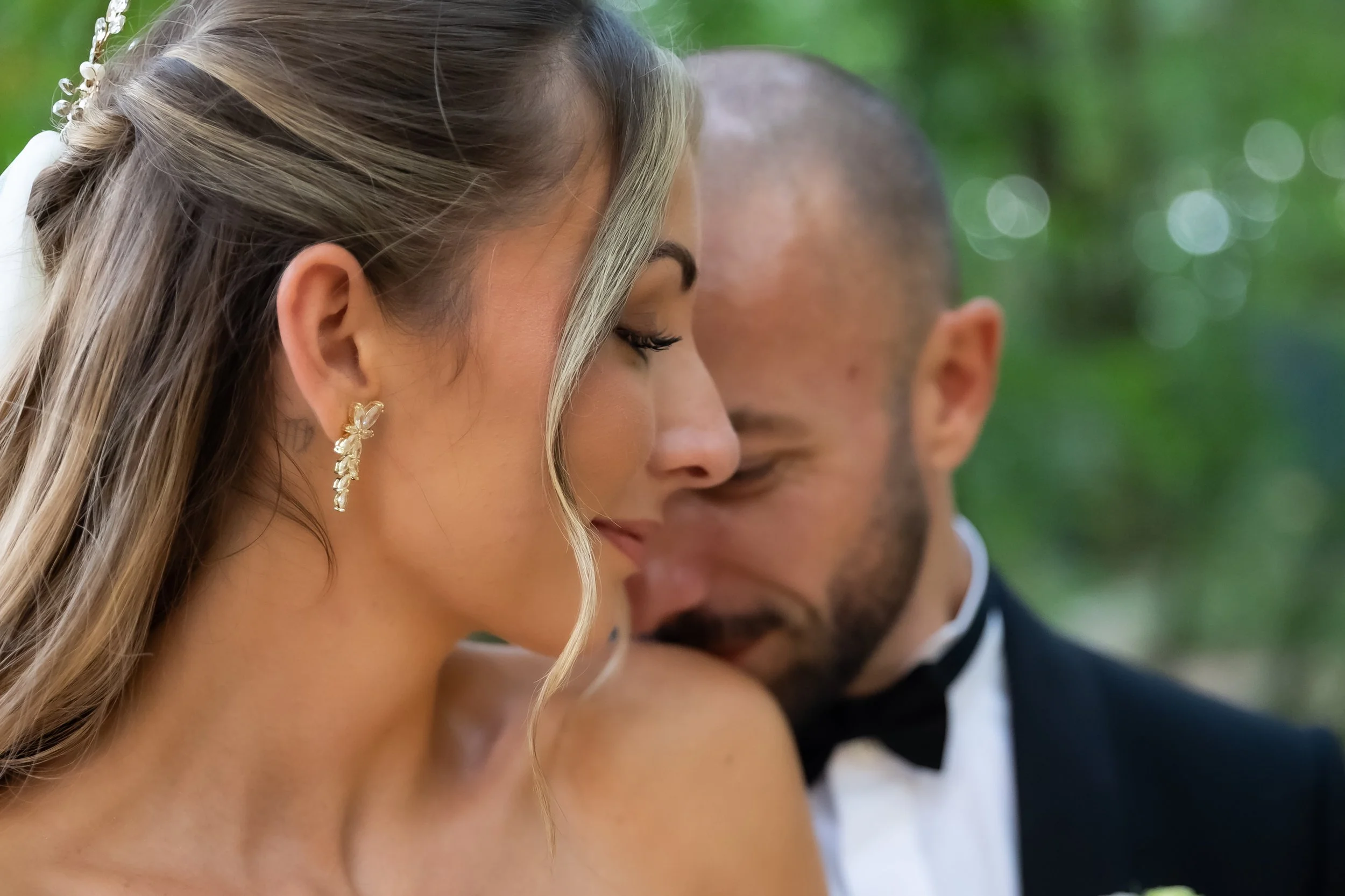 Close-up of a woman with blonde hair styled in waves, wearing gold floral earrings, and a man in a tuxedo with a bow tie, both with eyes closed and their foreheads touching, outdoors with blurred green background.