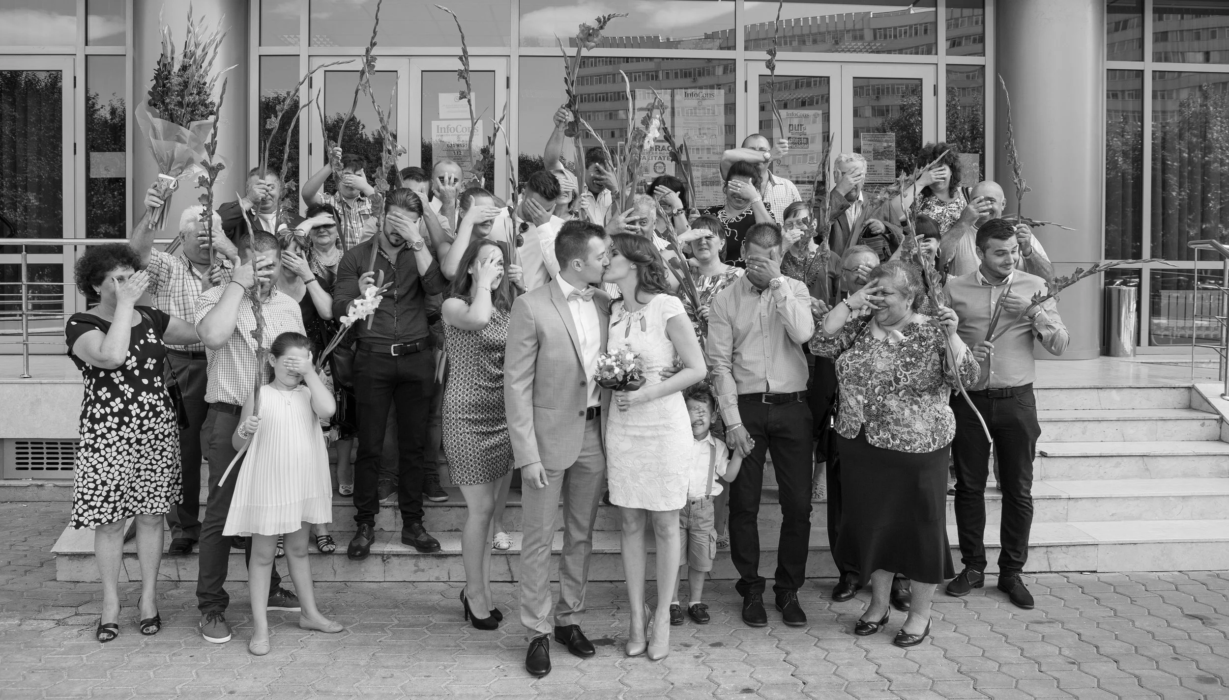 A group of people gathered outside a building, with some holding flower stems. In the center, a couple appears to be kissing or about to kiss, with the woman holding a small bouquet of flowers. Most individuals are covering their eyes, possibly in a 