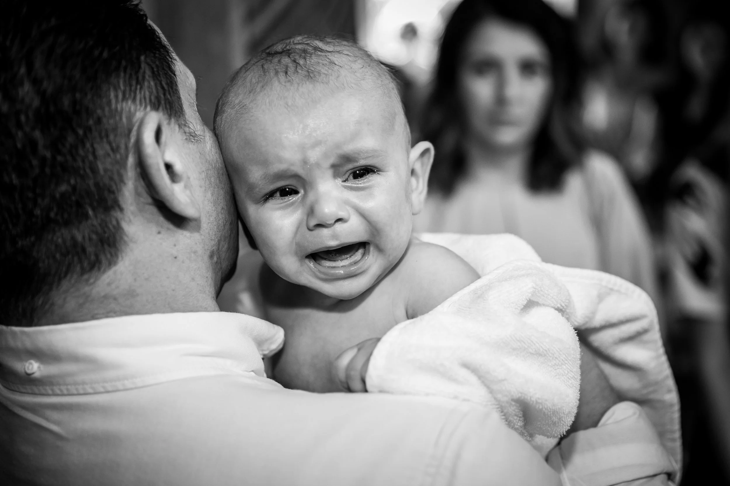 A person holding a crying baby in black and white, with a woman looking on in the background.
