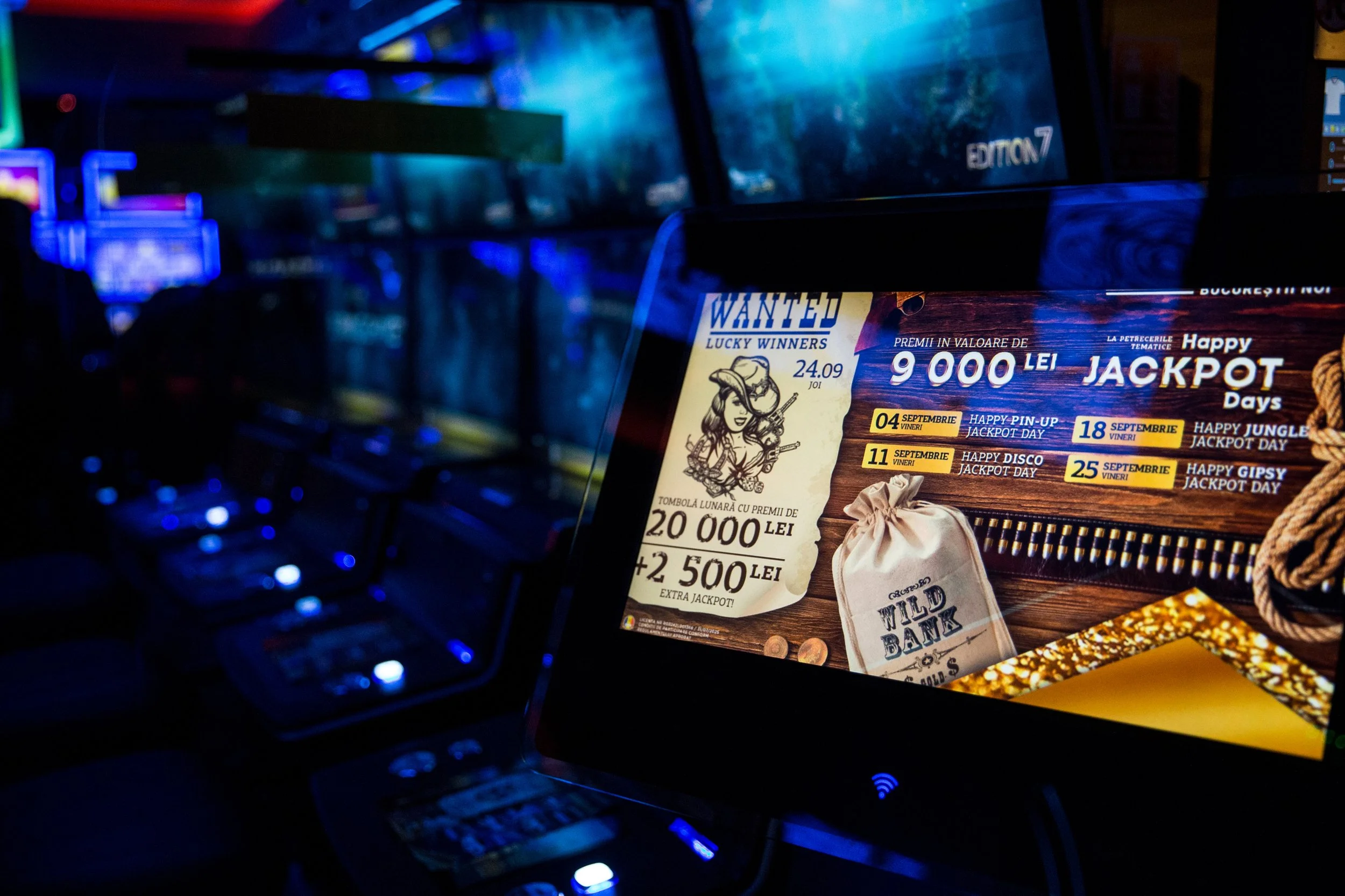 Close-up of a colorful casino or gaming center poster advertising jackpots and prizes, with a slot machine and a computer screen visible in a dimly lit environment with blue and purple lighting.