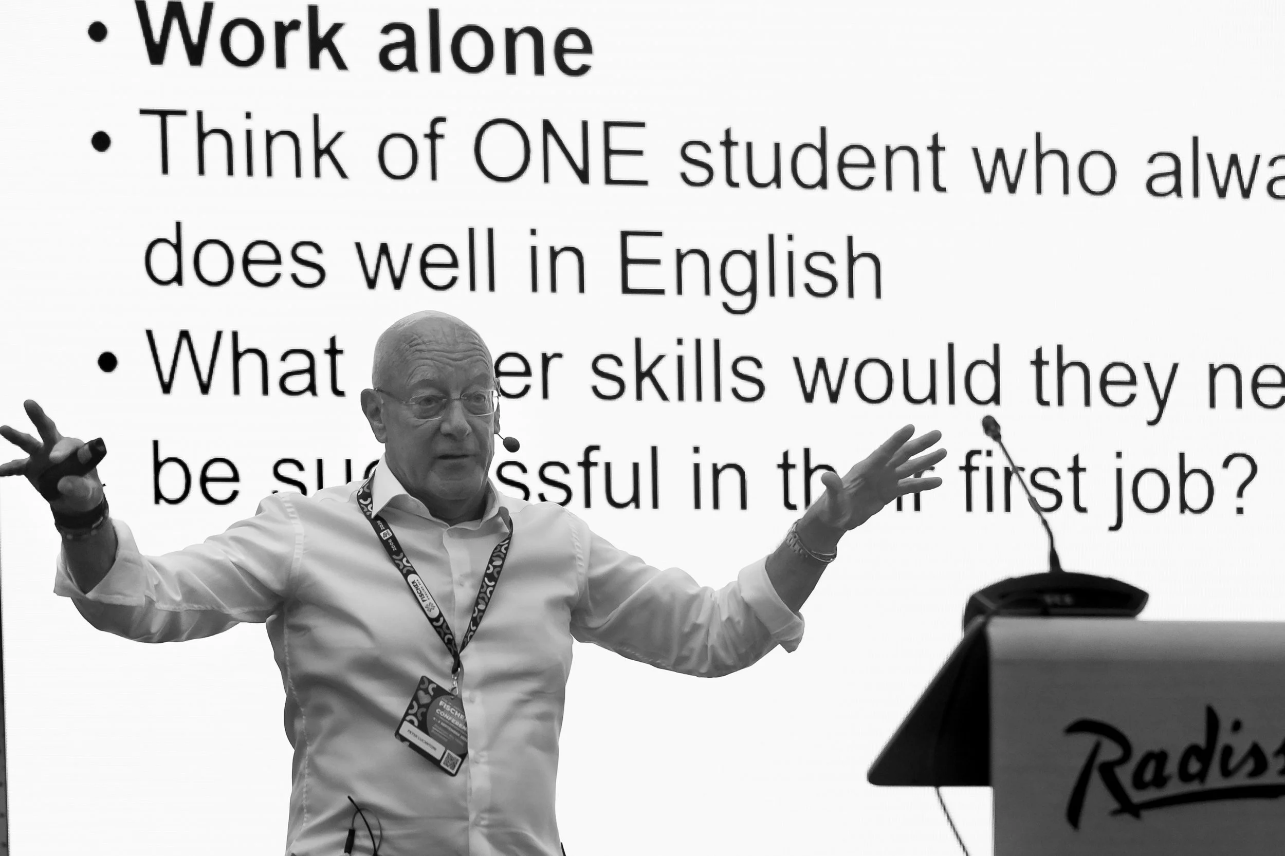 A man giving a presentation in front of a large screen displaying text about work and education, with his arms raised and wearing glasses, a white shirt, and a conference badge.