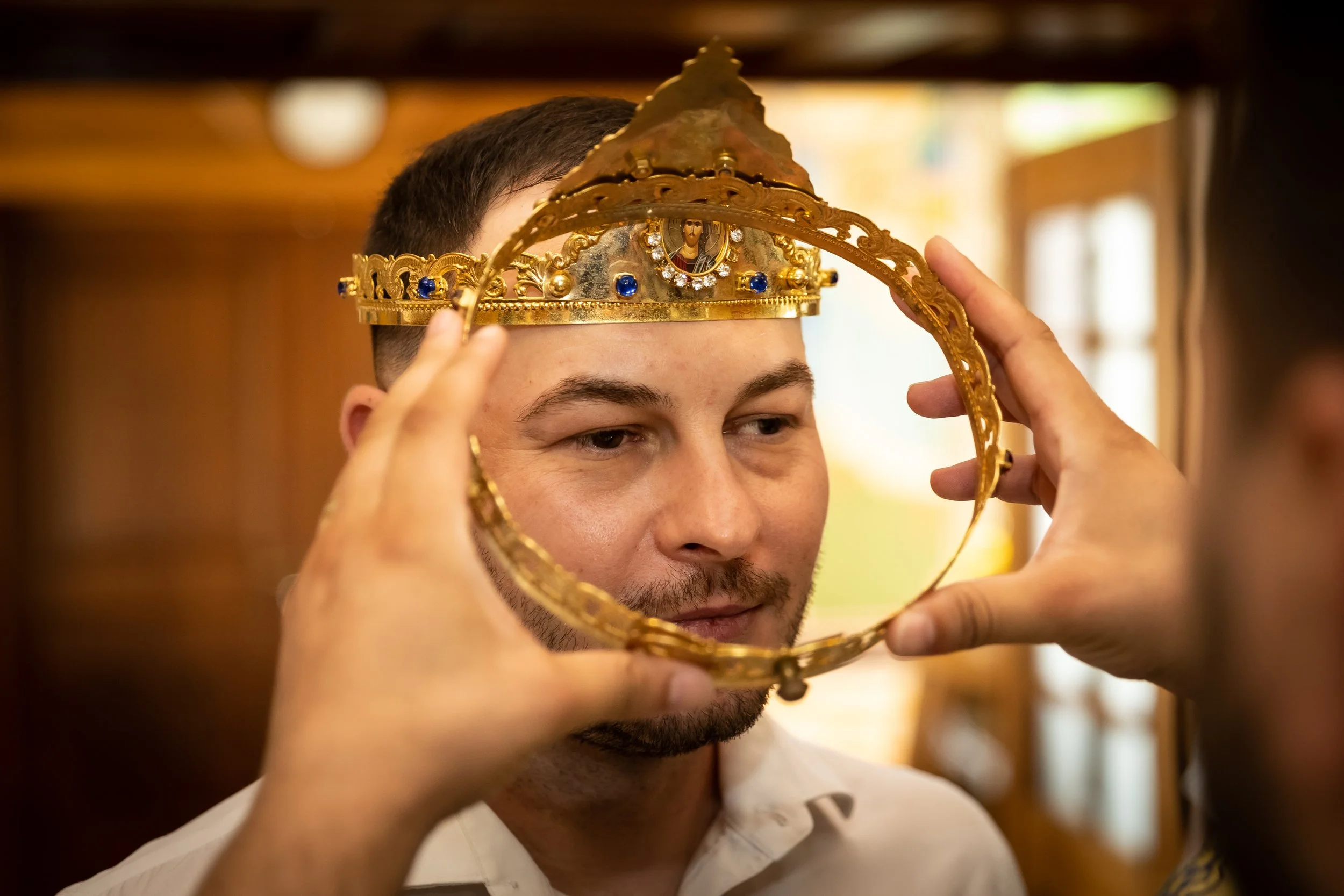 A young man with short dark hair and beard wearing a white shirt, holding a decorative gold crown in front of his face, with an image of a saint or religious figure on the crown.