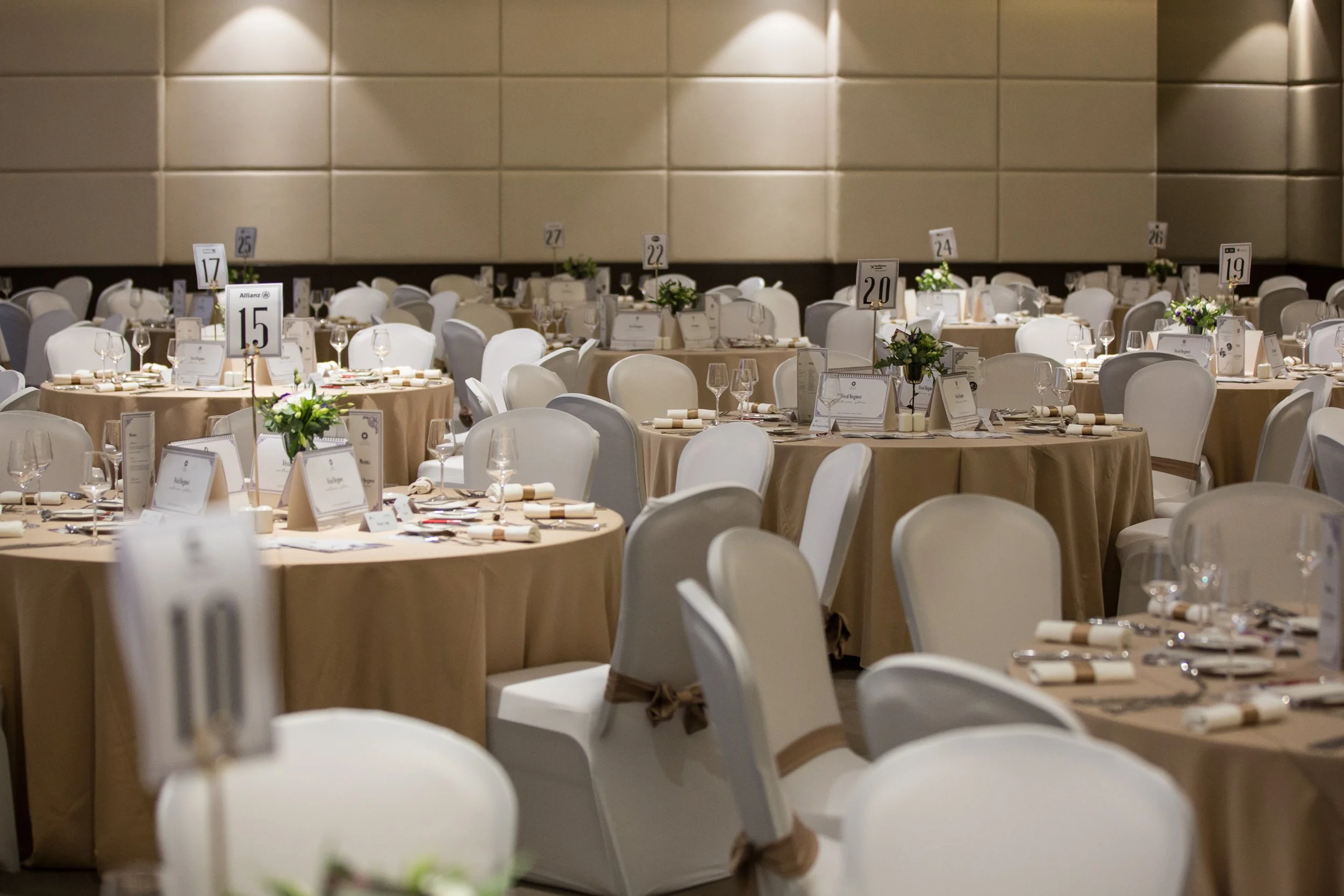 Multiple round banquet tables set with white chairs, wine glasses, napkins, place cards, and floral centerpieces in a large event hall with beige walls.