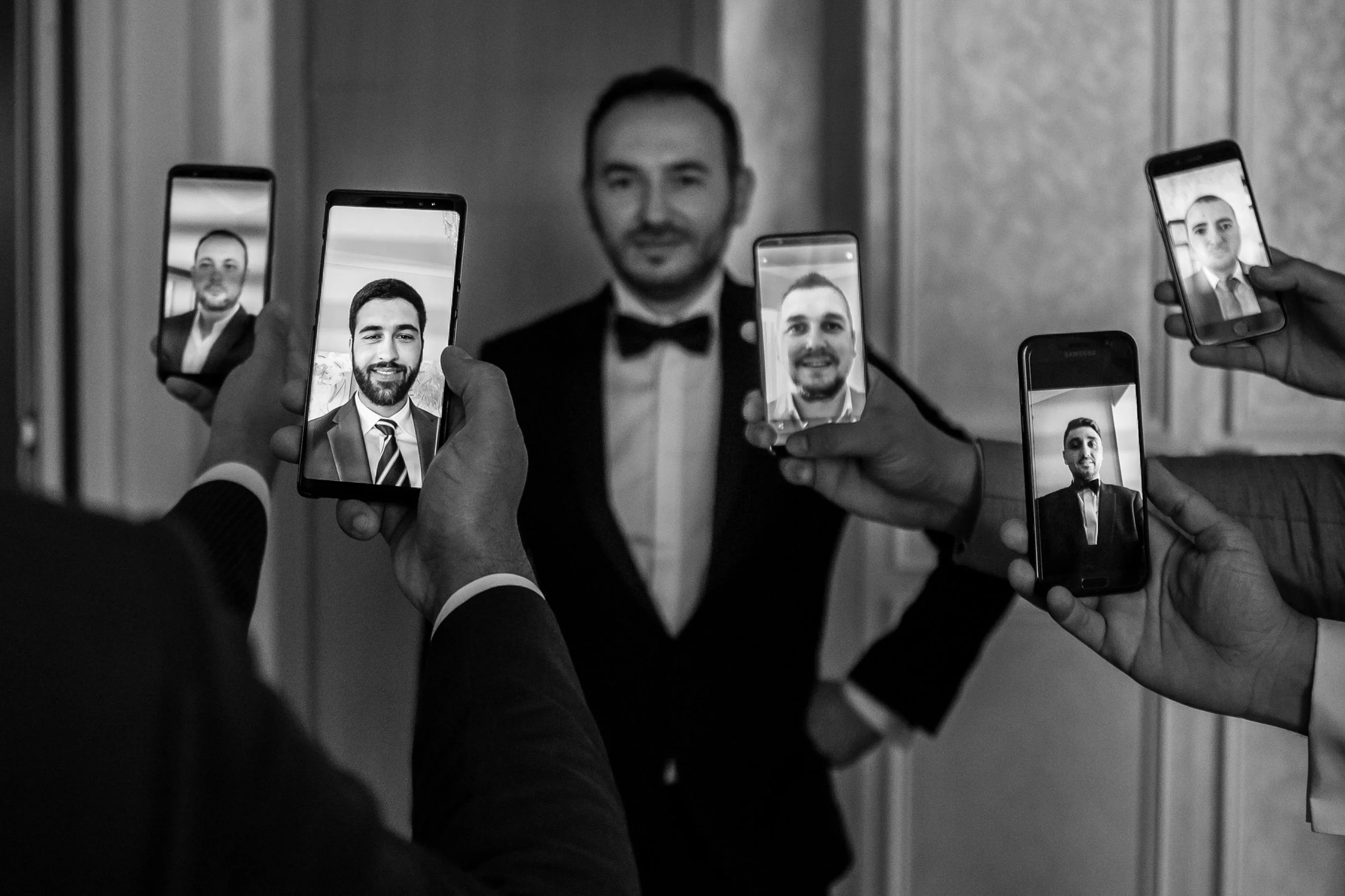 A man in a tuxedo taking a selfie with multiple smartphones held by different hands, capturing his image from various angles.