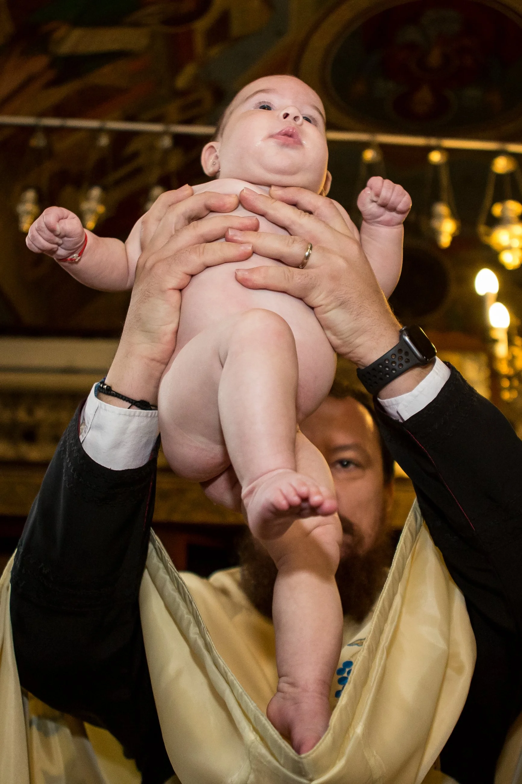 A man holding a baby in a yellow and black baby carrier inside a room with warm lighting and ornate ceiling details.