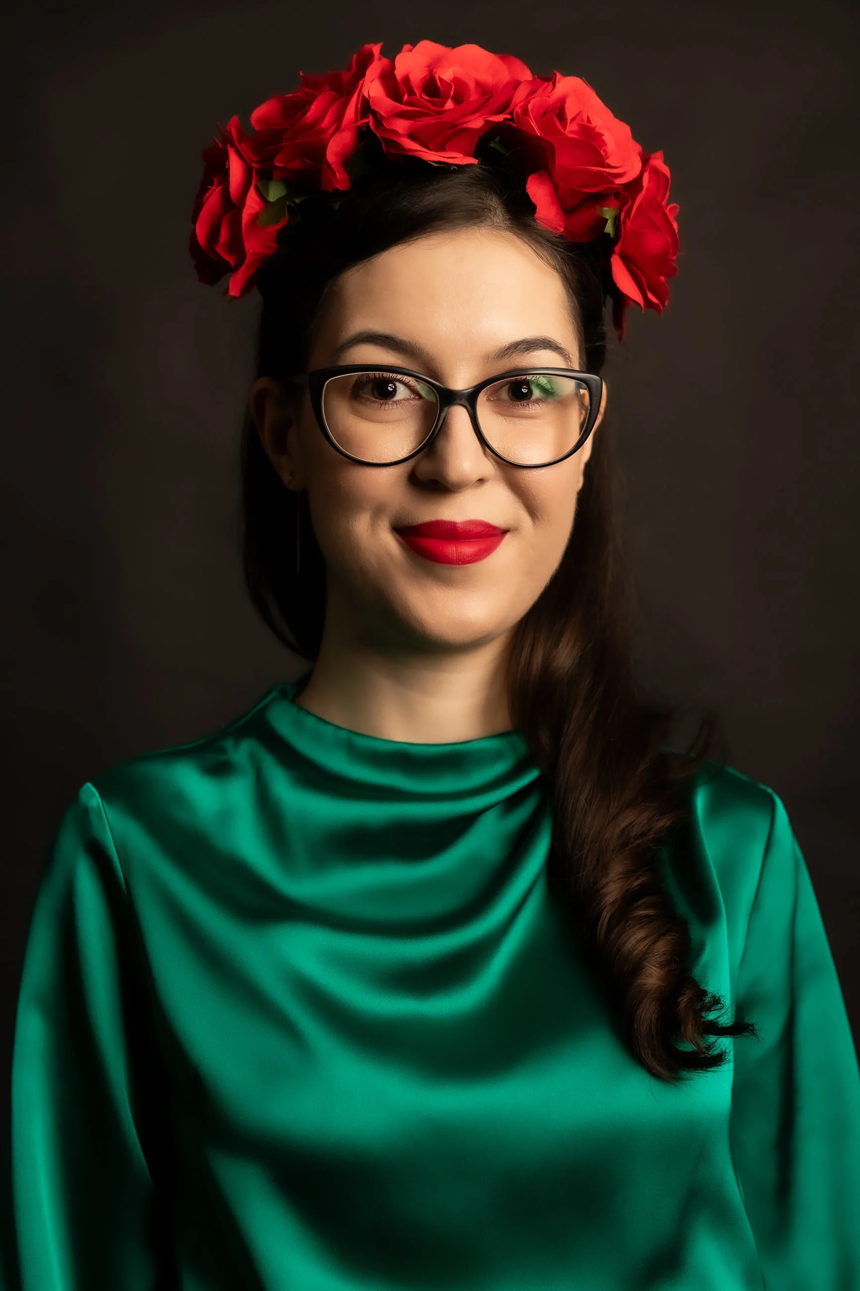 A woman with long brown hair wearing black glasses, red lipstick, a green satin dress, and a crown of red roses on her head, against a plain dark background.