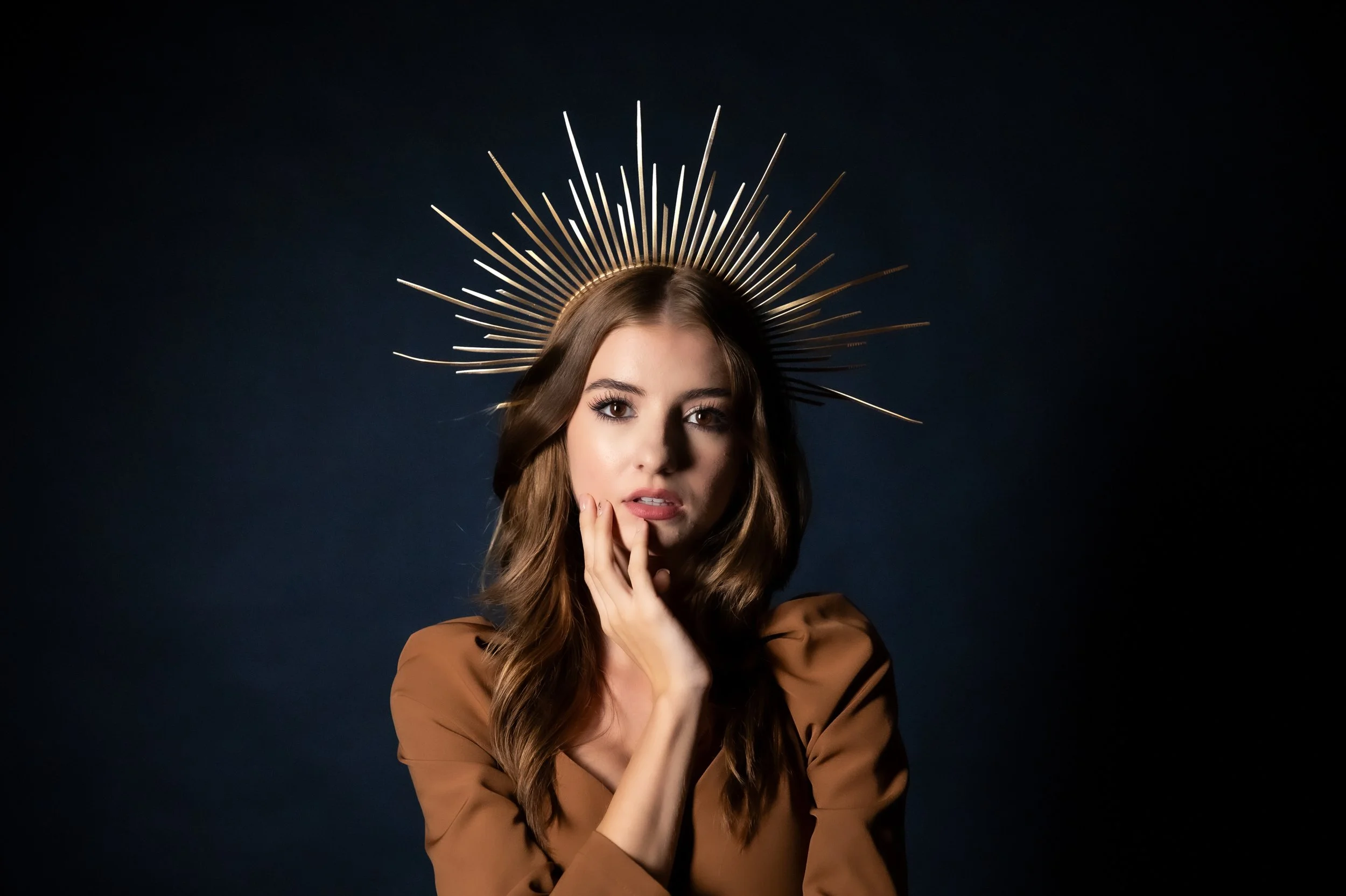 A woman with long wavy hair wearing a brown top and gold crown with radiating spikes, posing against a dark background.