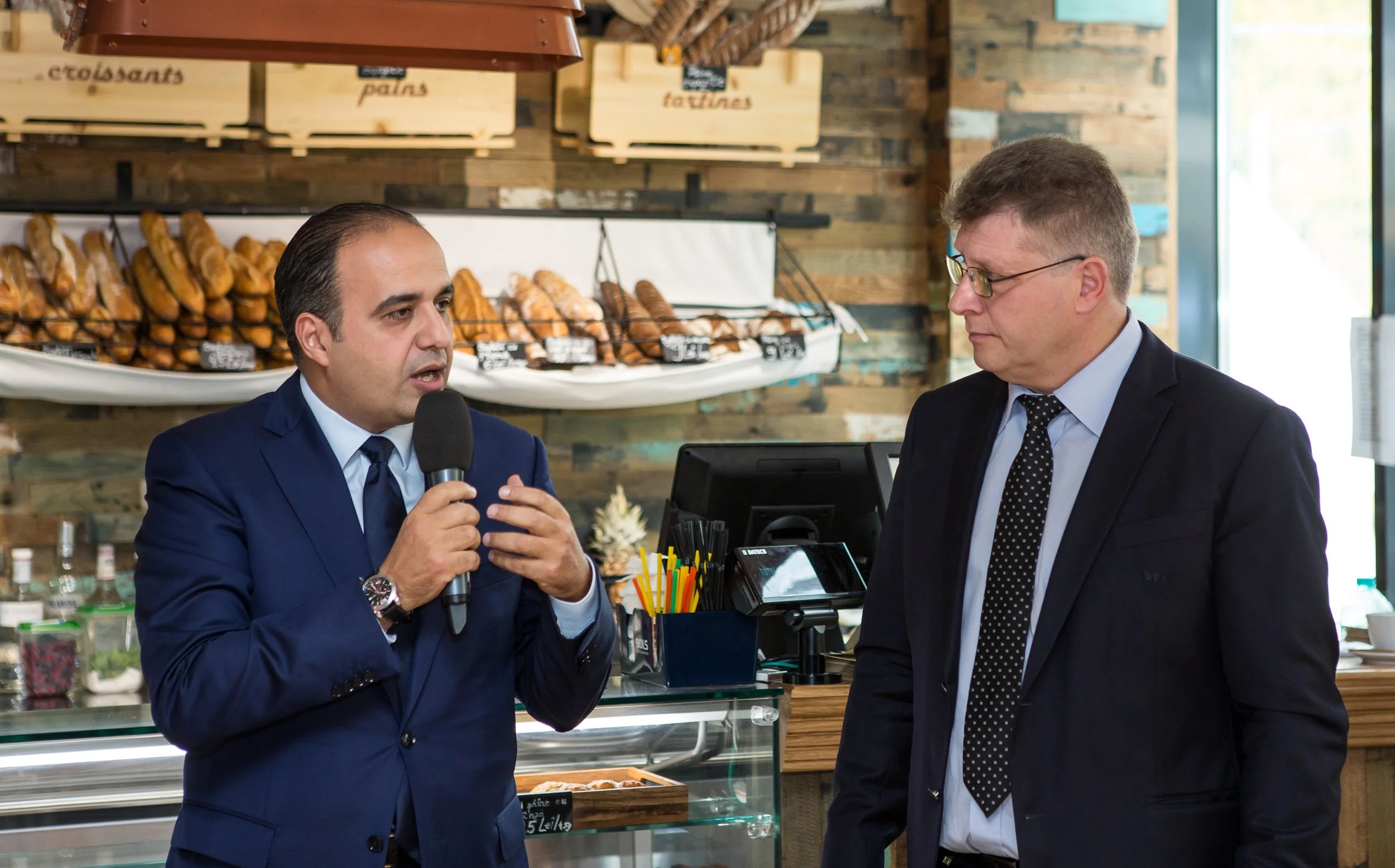 Two men in suits having a conversation inside a bakery or cafe, with pastries displayed in the background.